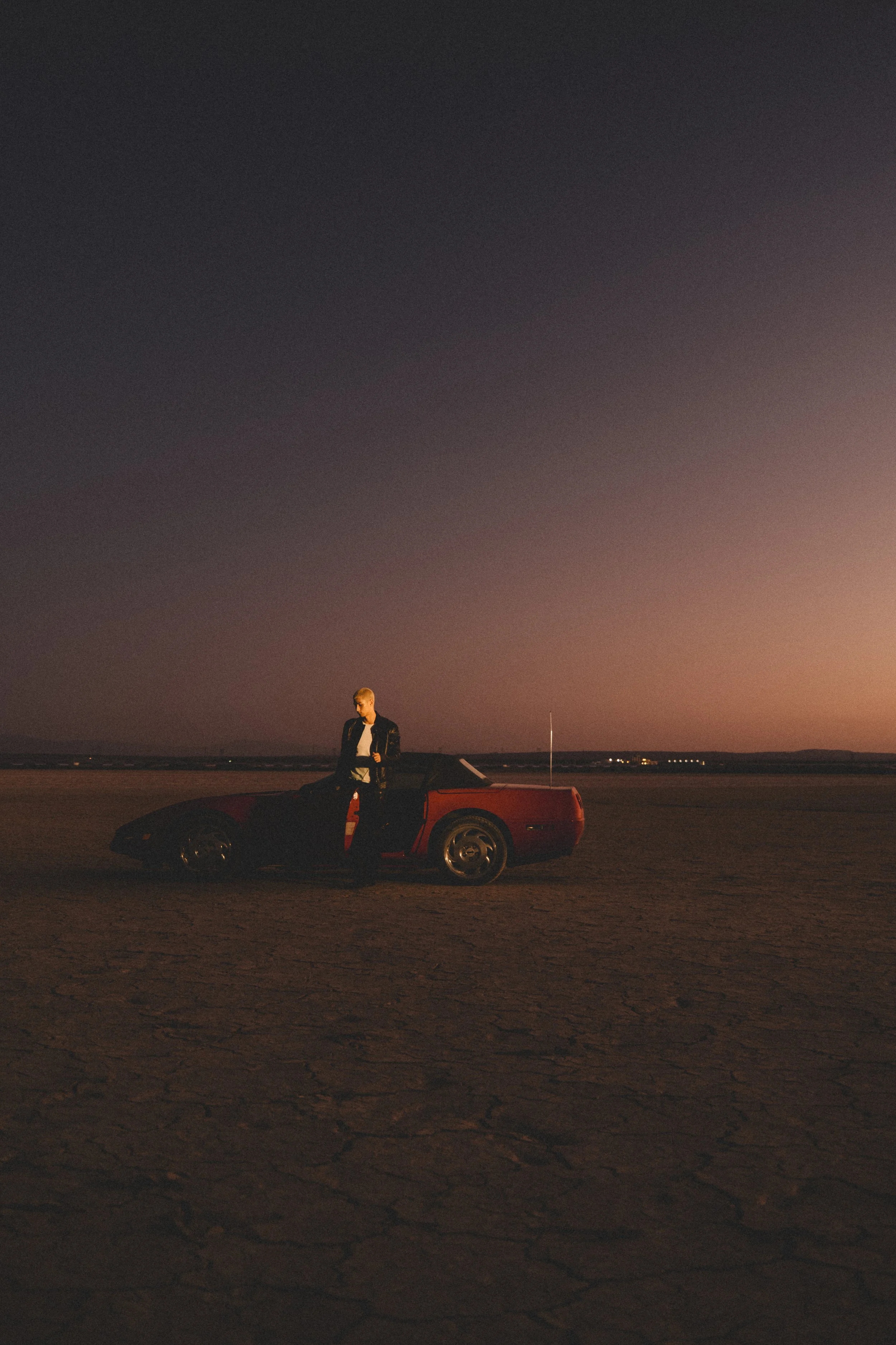 A person stands next to a red sports car on a dry, cracked landscape during sunset or sunrise, with a darkening sky in the background.