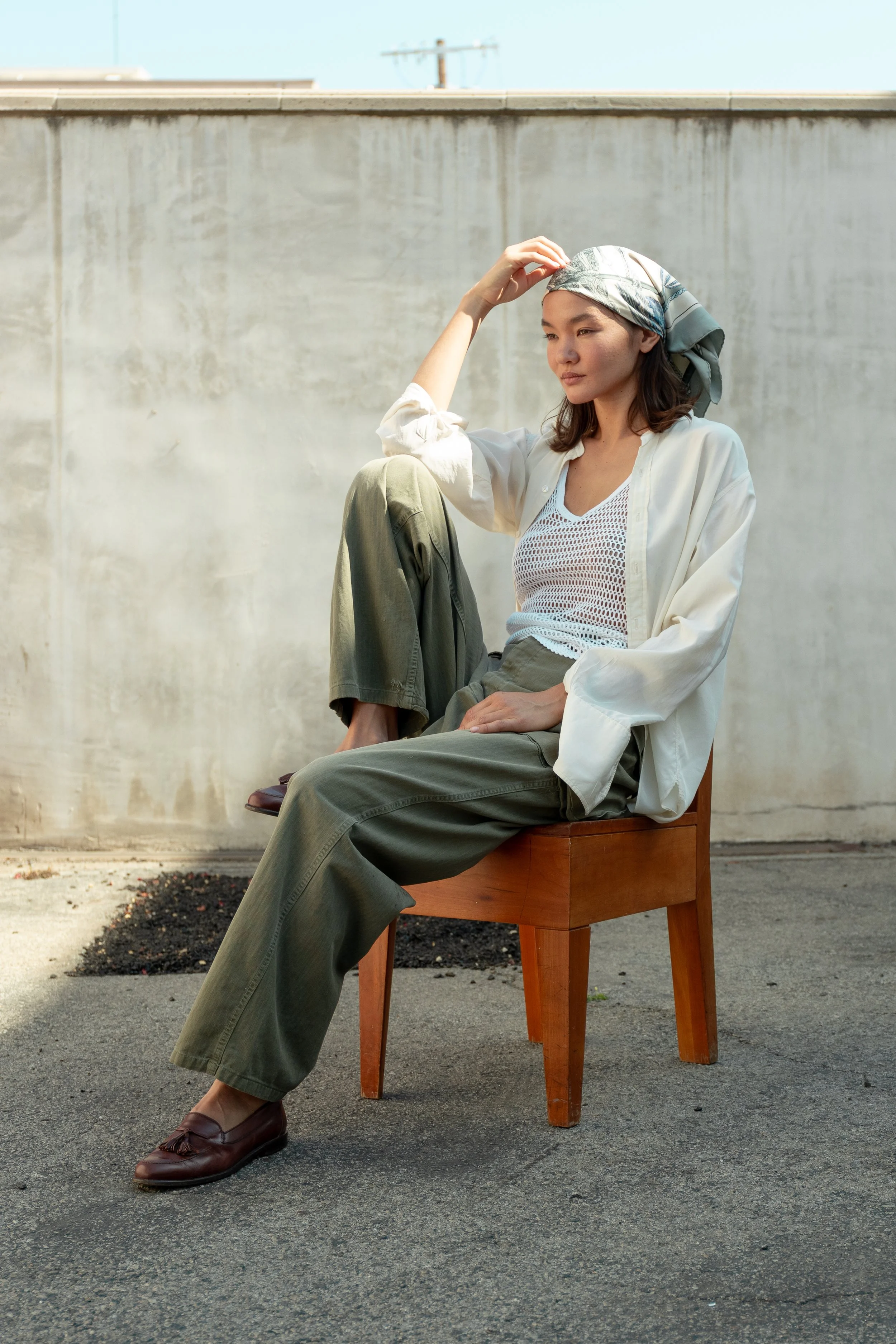 A woman sitting on a wooden chair outdoors against a concrete wall, wearing a headscarf, white open shirt over a mesh top, olive-green pants, and brown loafers, looking contemplative.