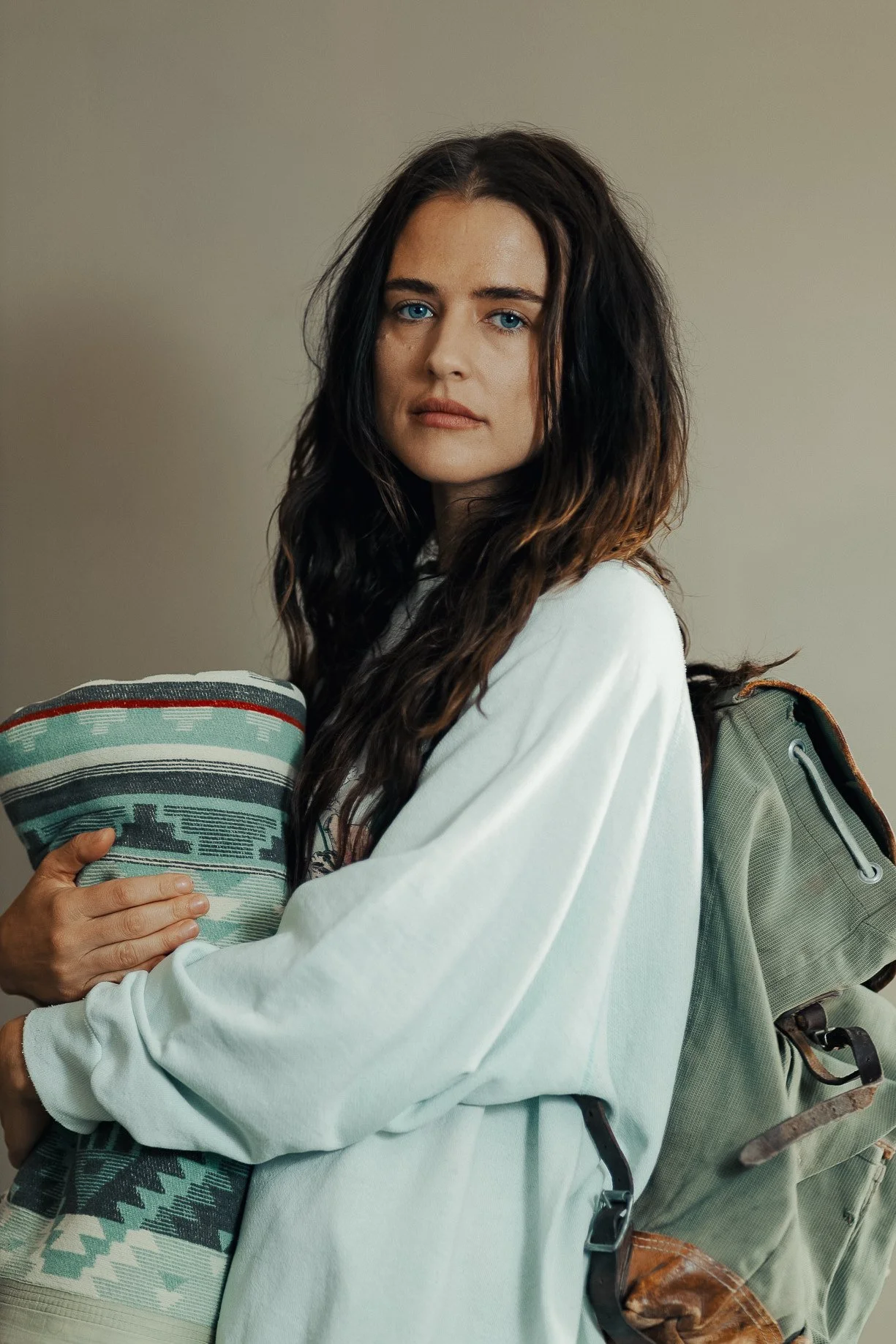 A young woman with long wavy brown hair and blue eyes wearing a white hoodie, holding a patterned fabric item, standing with a backpack on her shoulder against a plain background.