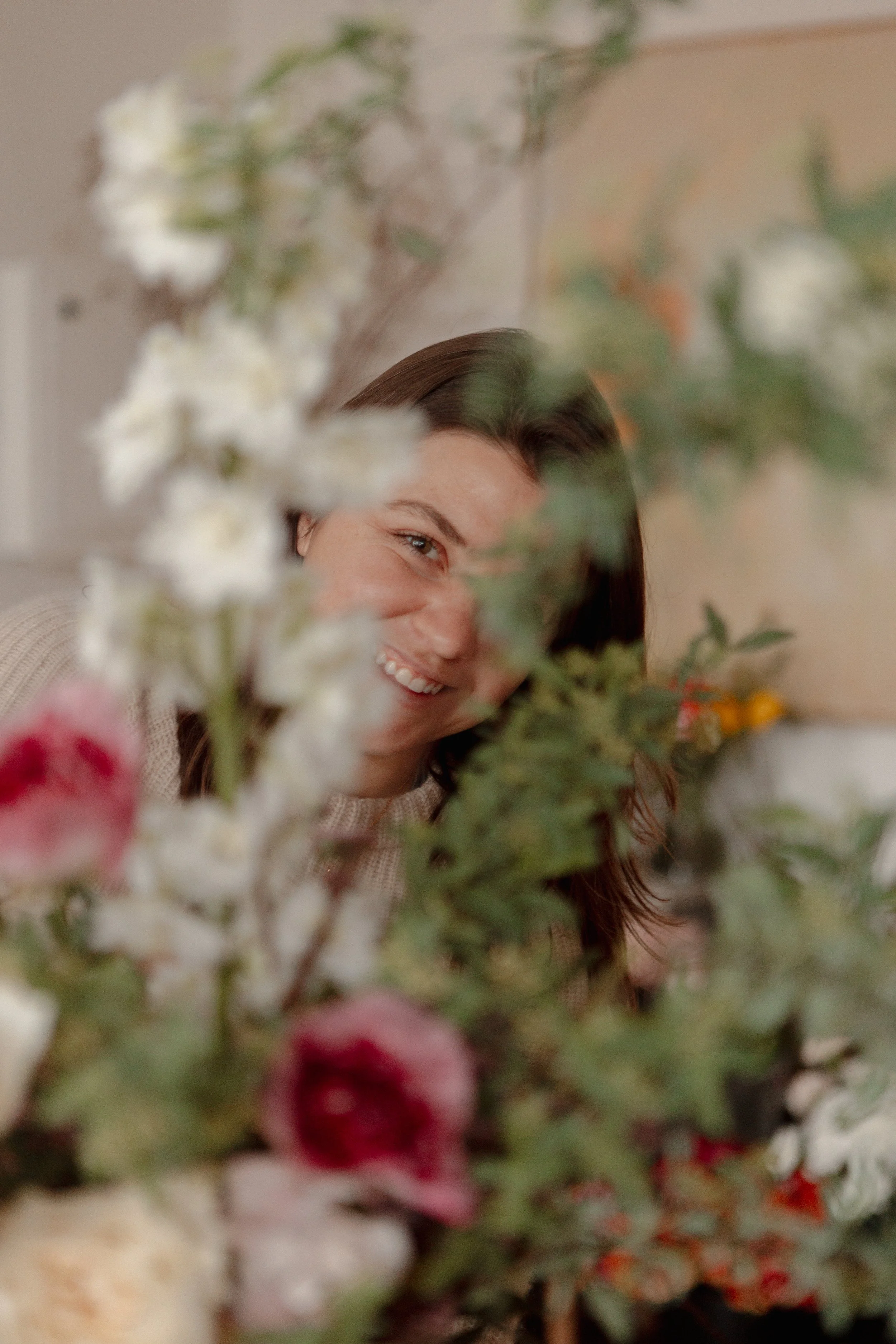 A woman smiling behind a bouquet of flowers, partially obscured by blurred flowers in the foreground.