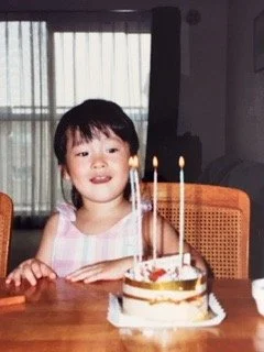 Young girl with dark hair and bangs celebrating birthday with a cake and lit candles indoors.