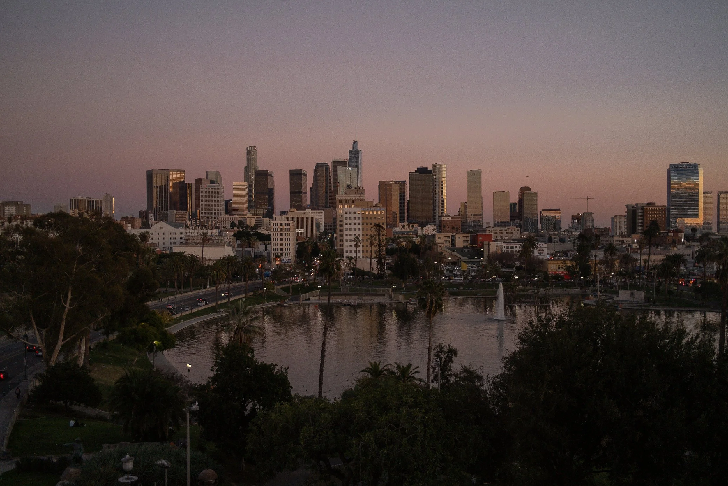 Skyline of Los Angeles with tall skyscrapers at sunset, view over a park with a pond and fountain, trees, and city streets.