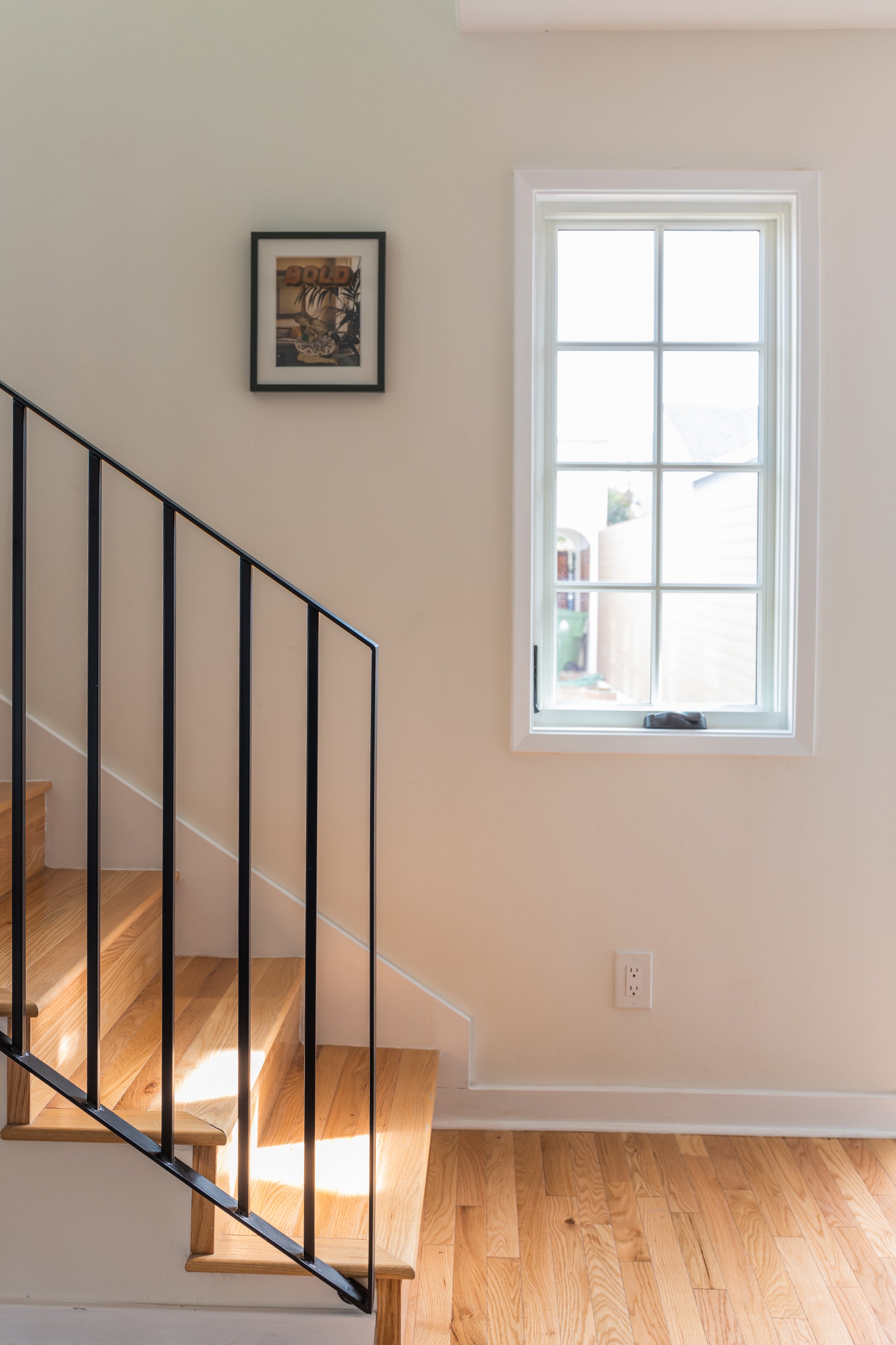 Interior of a house showing a staircase with wooden steps and black railing, a window with white trim, a framed picture on the wall, and a wooden floor.