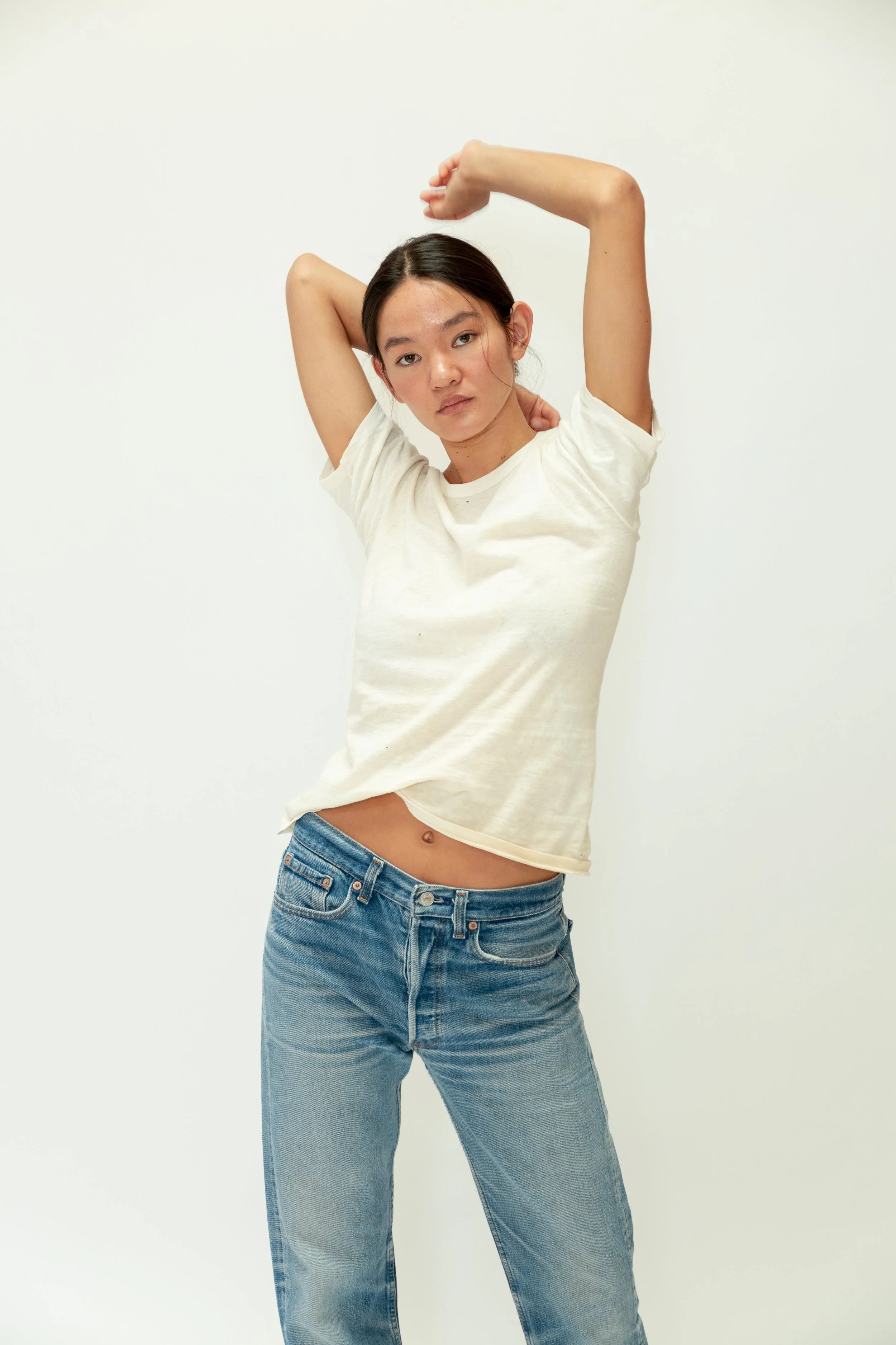 A young woman with dark hair and light skin posing with one arm raised and her hand touching her head, wearing a white t-shirt and blue jeans, standing against a plain white background.