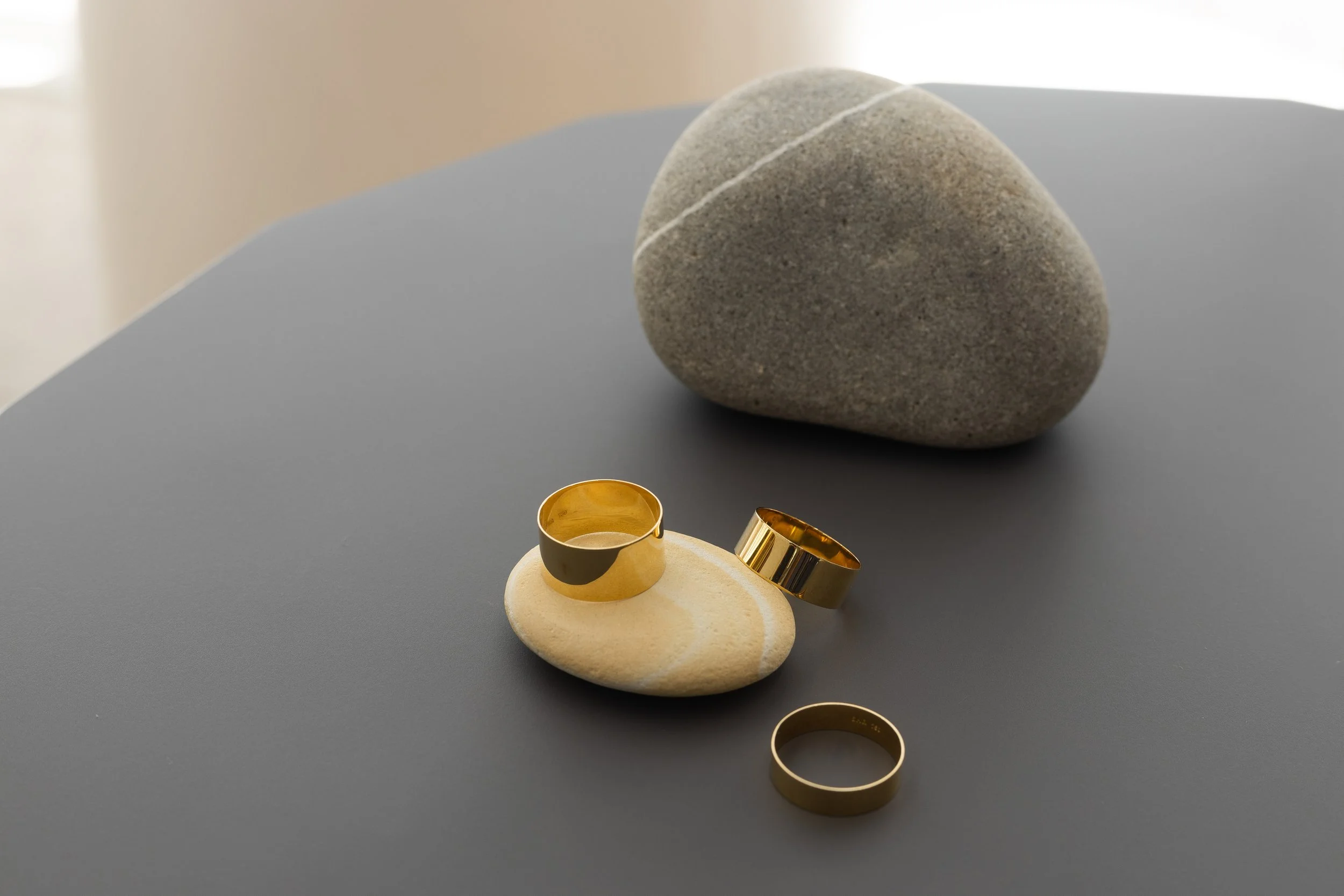 Three gold rings placed on a light-colored shell, with a large gray stone in the background on a dark gray surface.