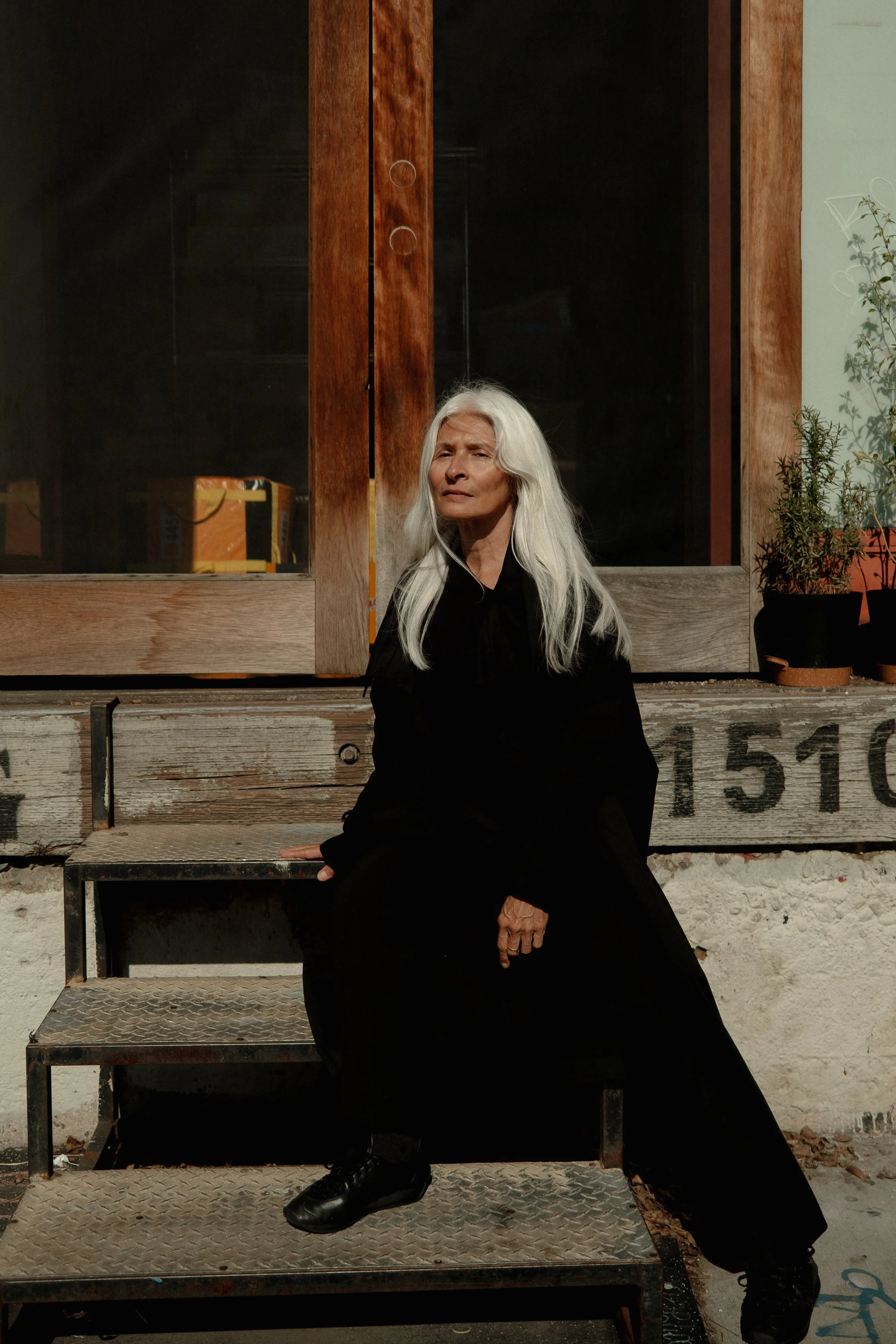 An elderly woman with long white hair, dressed in black, sitting on a staircase outside a building with a wooden door and window, with potted plants nearby, in natural sunlight.
