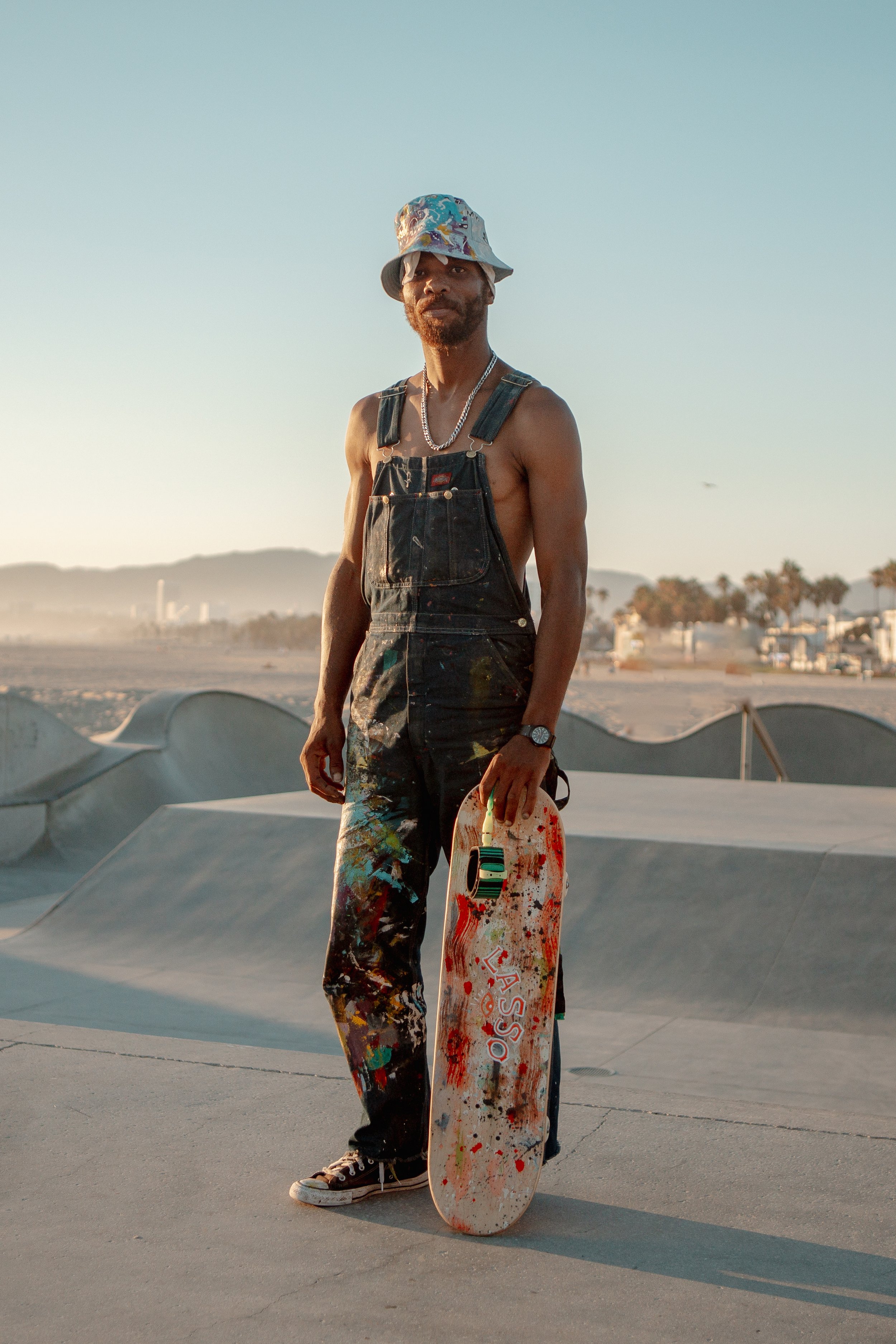 A shirtless man wearing overalls, sneakers, and a colorful bucket hat holding a skateboard at a skate park during sunset.