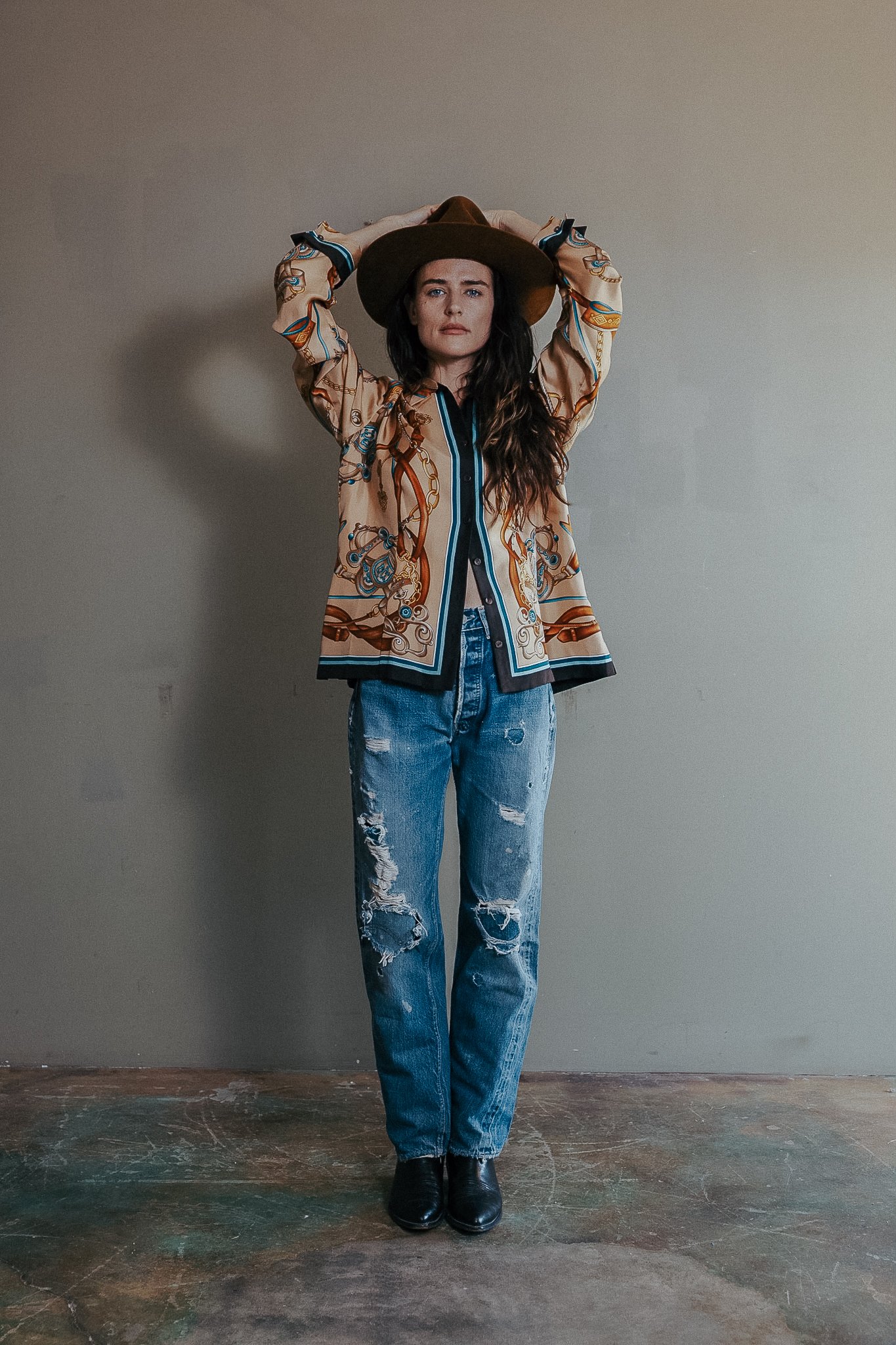 A woman standing against a plain wall, wearing a patterned jacket, distressed jeans, black boots, and a wide-brimmed hat, holding her hat with her hands.