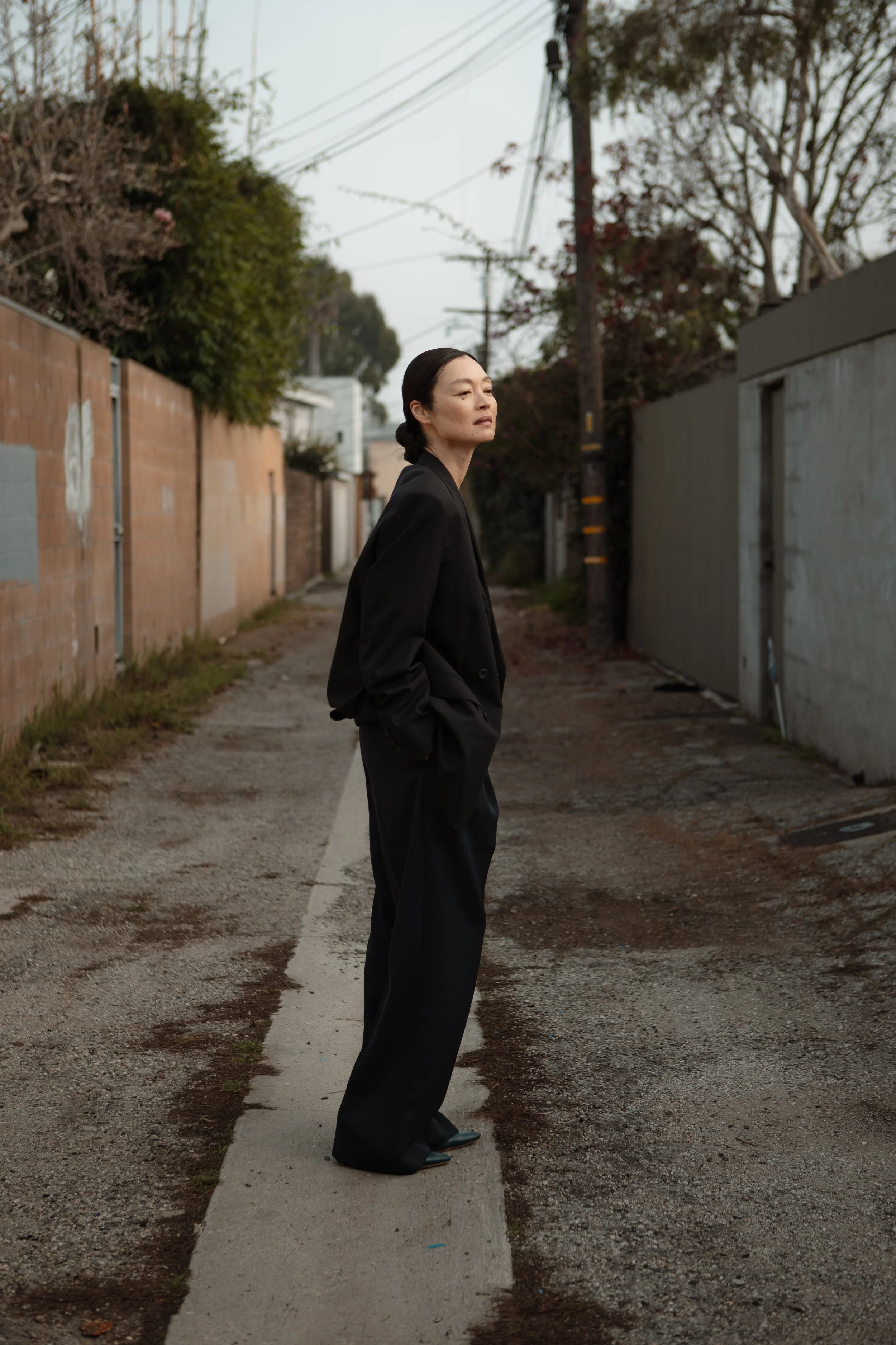 A woman in black clothing standing on a narrow dirt path in an alleyway with a neutral expression, surrounded by a brick wall on one side and a concrete wall on the other, with overhead power lines and trees in the background.