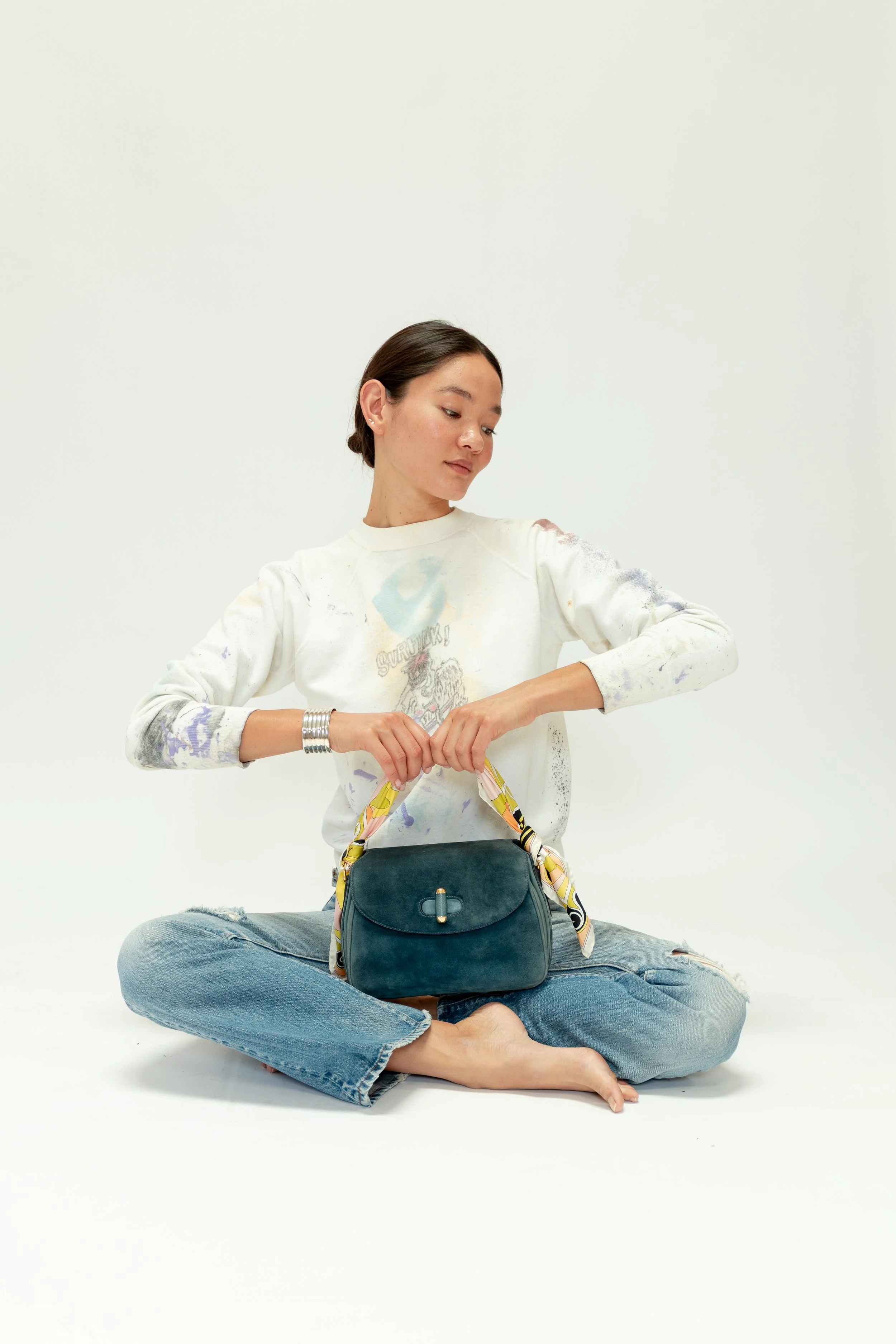 A young woman sitting cross-legged on the floor, wearing a white sweatshirt with colorful paint splatters and printed graphics, holding a teal handbag with a yellow scarf tied to its handle, against a plain white background.