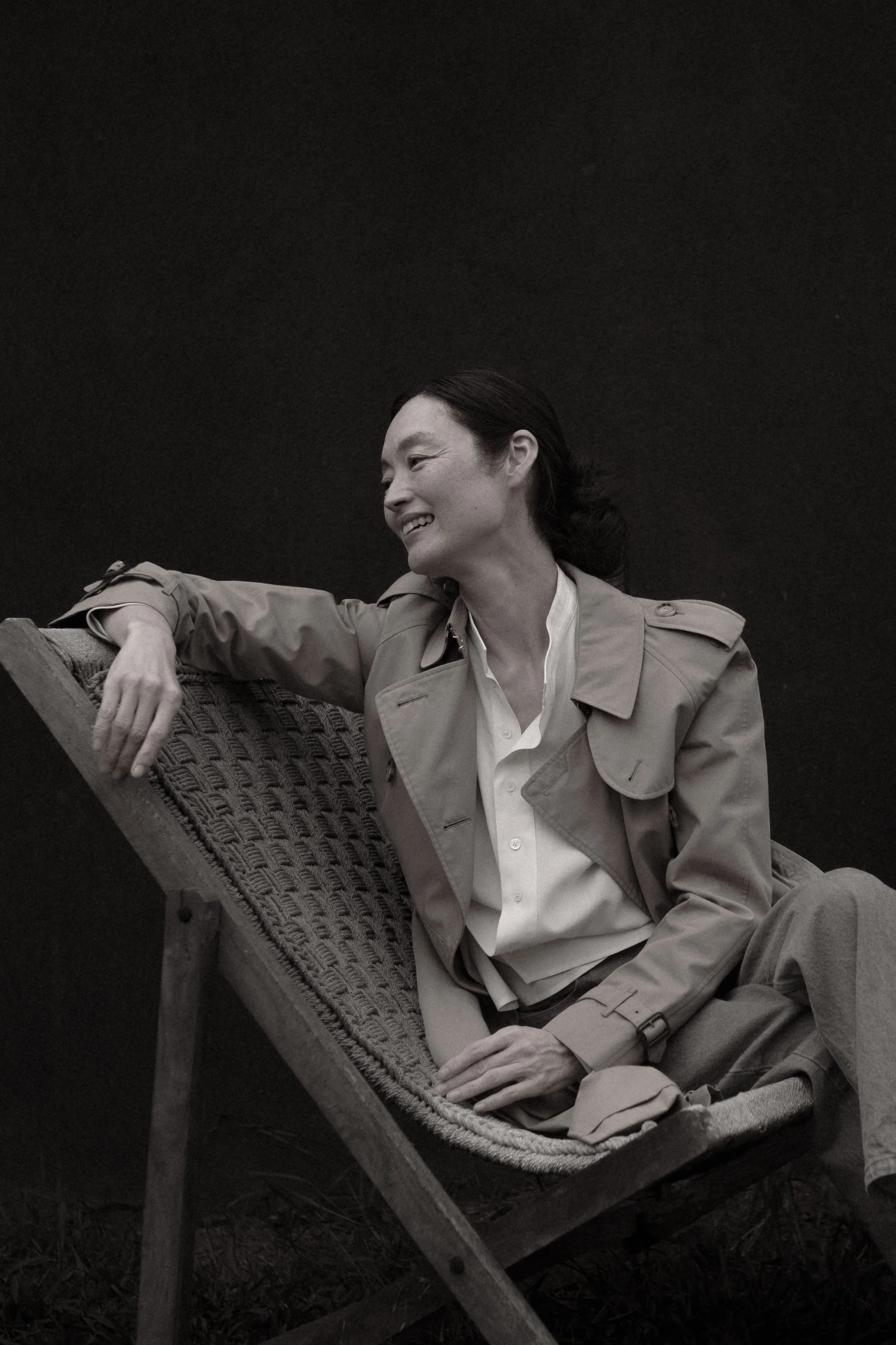 A woman reclining on a wooden lounge chair, smiling and looking to the side, wearing a trench coat and dress shirt, against a plain dark background.