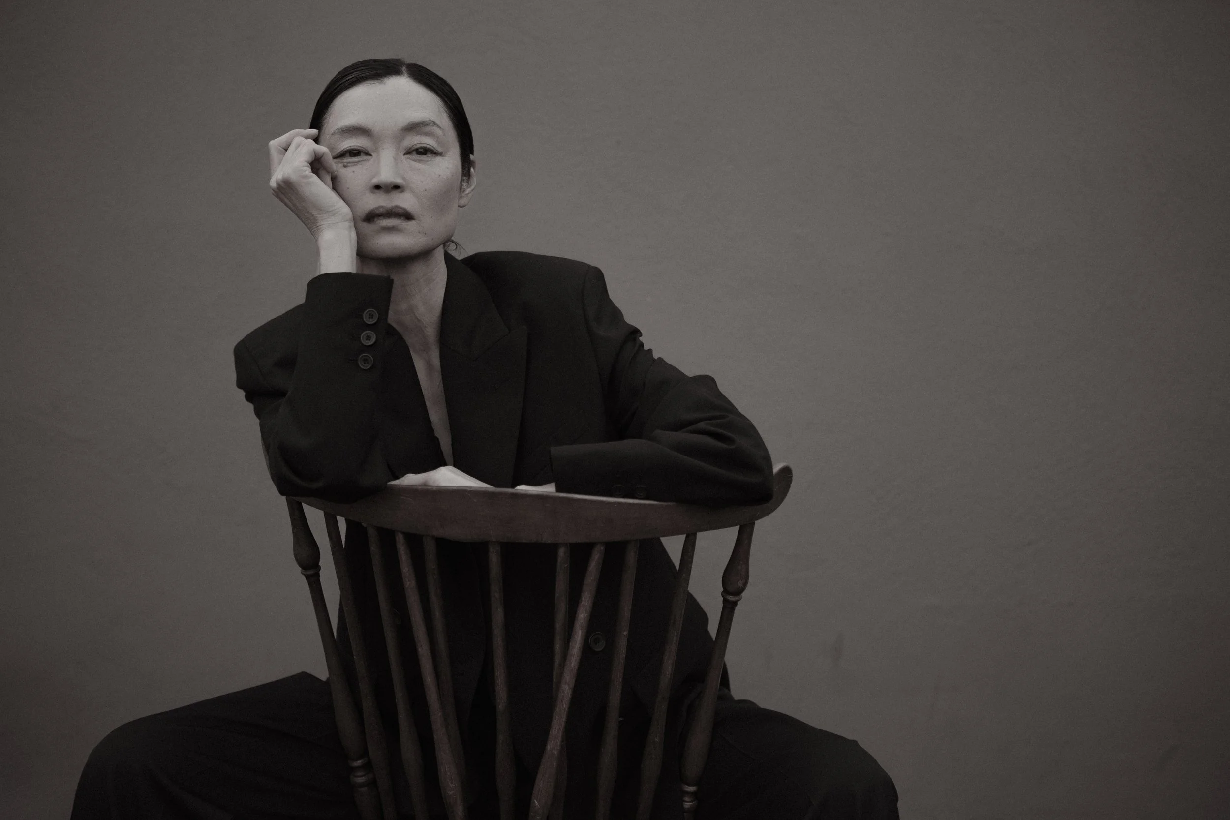 Black and white portrait of a woman sitting on a wooden chair with a high back, wearing a dark blazer, resting her right elbow on the back of the chair, with her right hand gently touching her face, against a plain background.