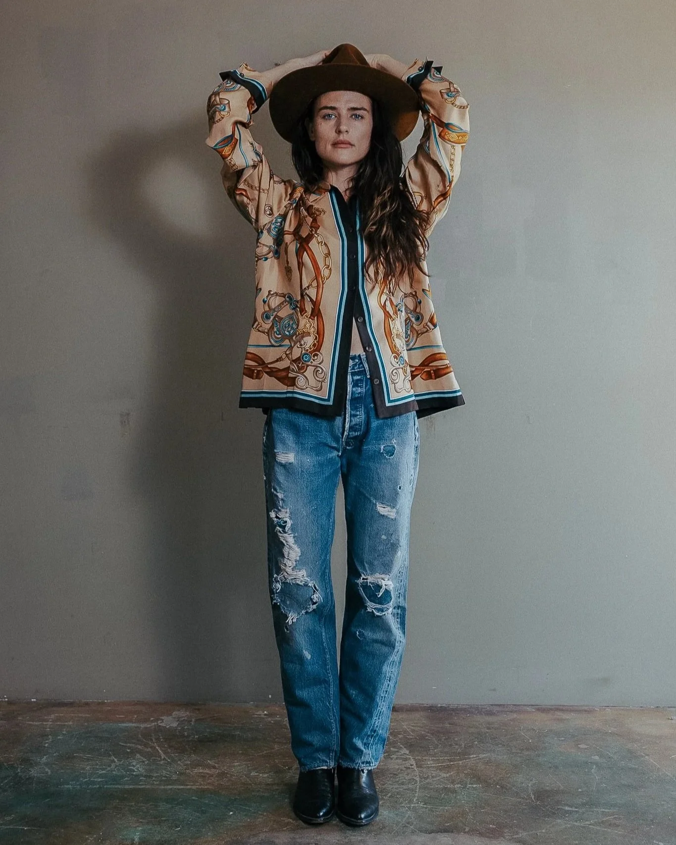 A woman with long dark hair wearing a large brown hat, a beige patterned blouse, ripped blue jeans, and black boots, standing against a plain wall.