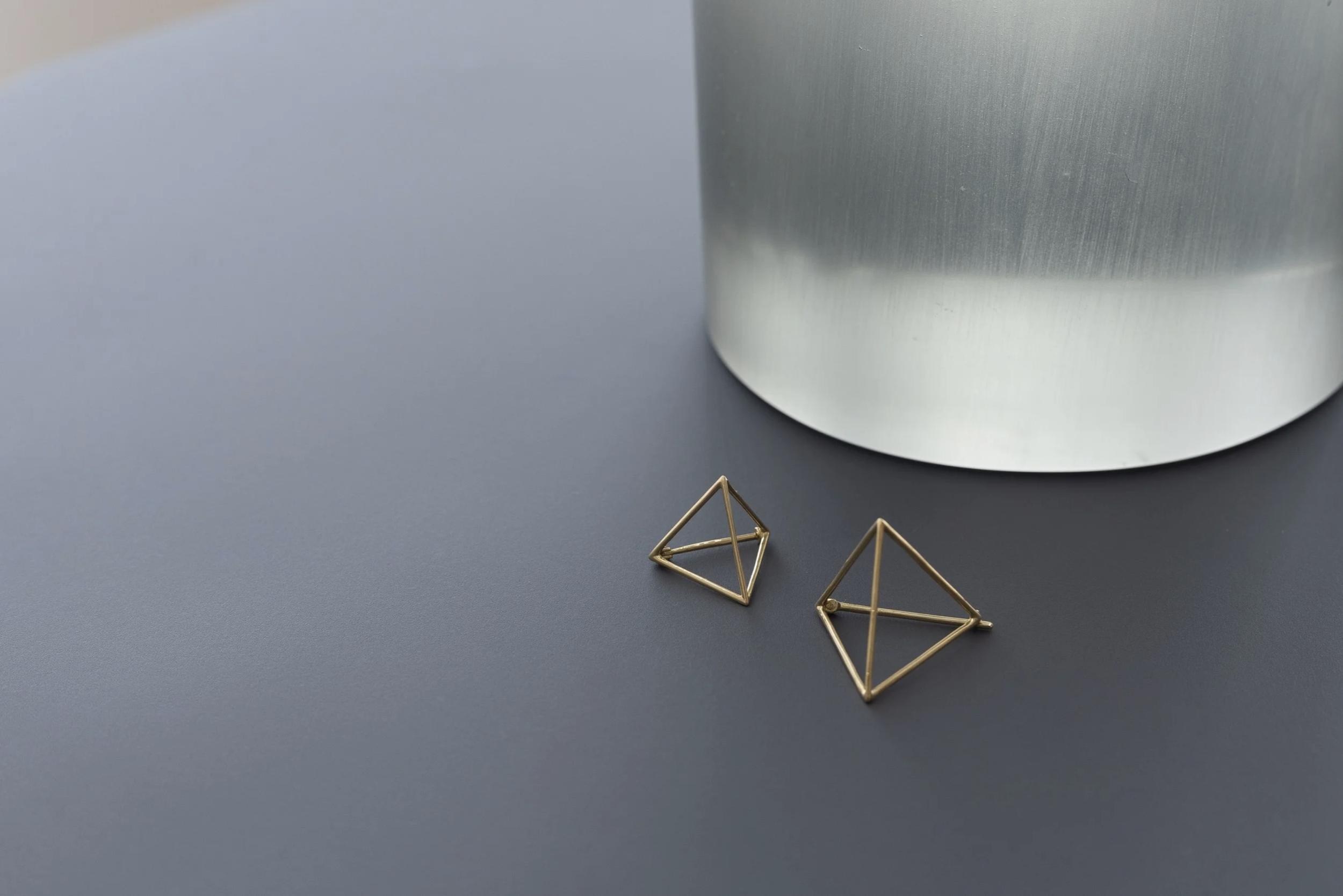 Pair of geometric gold earrings with triangular and tetrahedral shapes on a gray surface, with a blurred metallic object in the background.