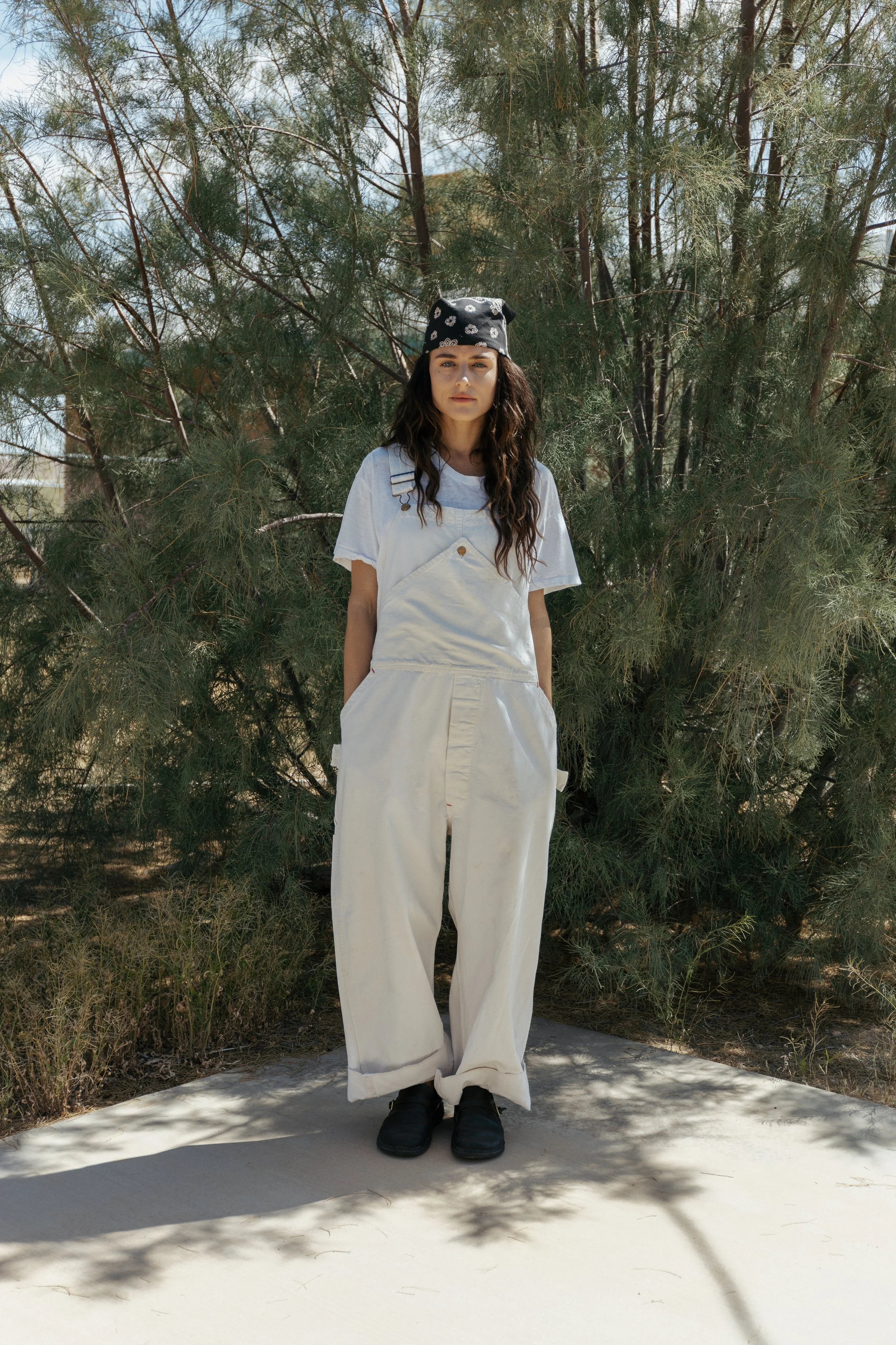 A woman standing outdoors in front of a green tree, wearing a black bandana, a white t-shirt, white overalls, and black shoes.