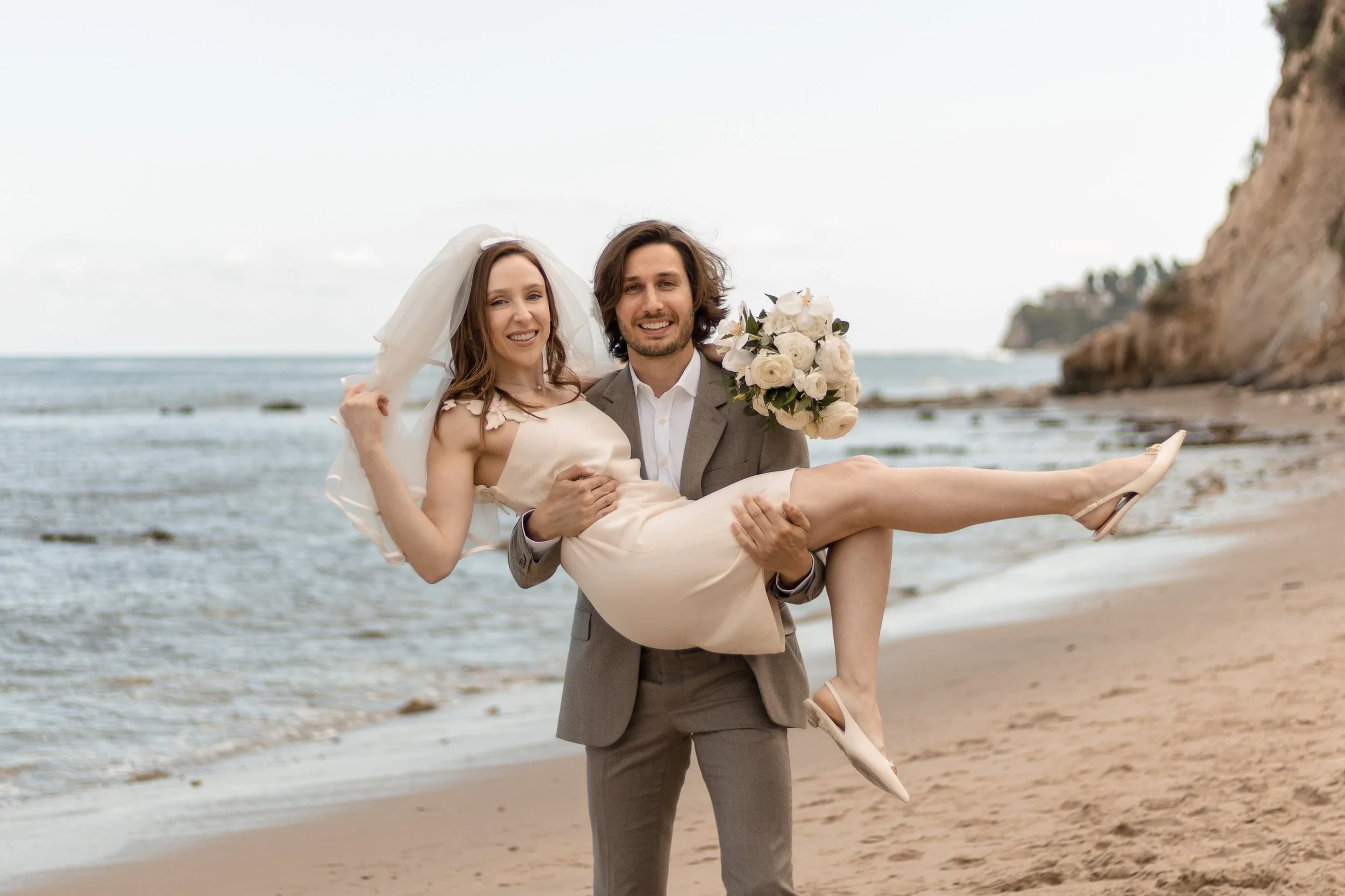 A smiling groom holding a bride in a wedding dress with a bouquet of white roses, on a beach with cliffs in the background.