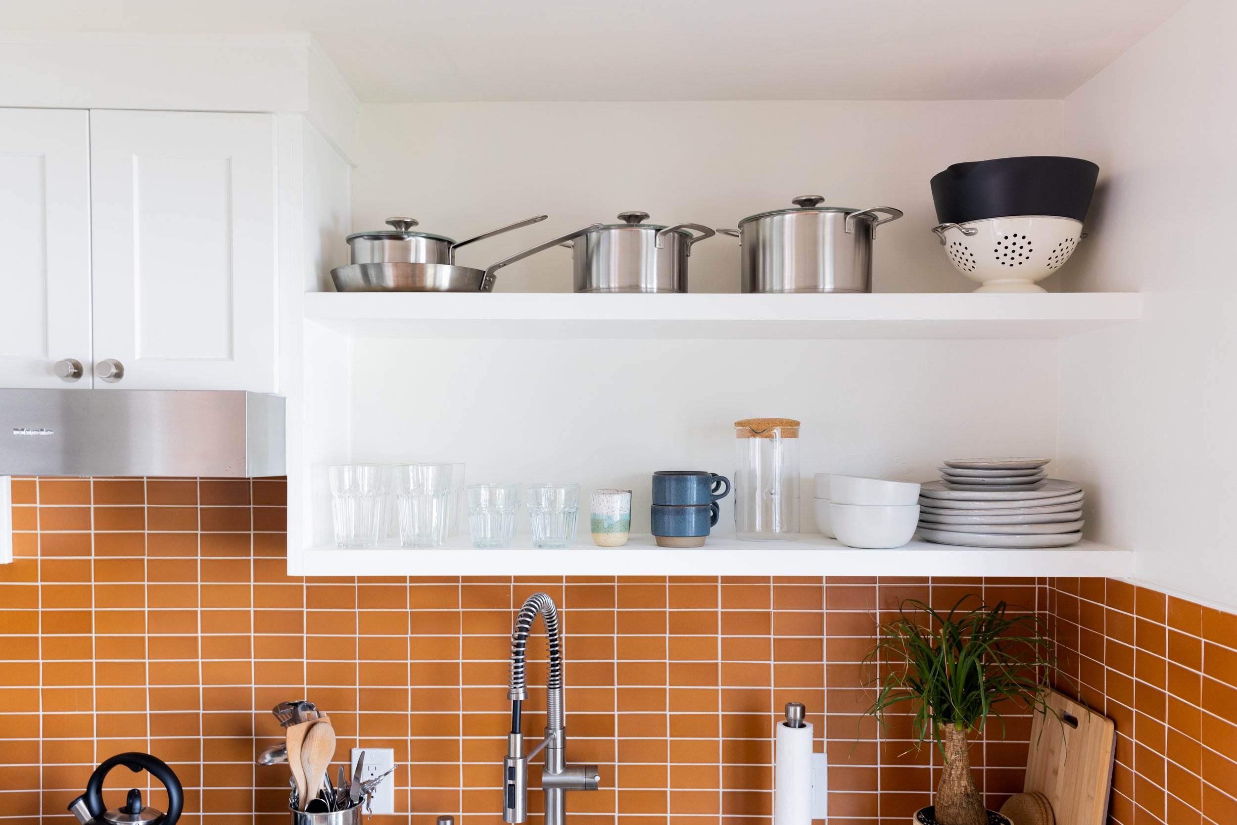 Open kitchen shelves with stainless steel pots and a colander, white bowls, glassware, and a decorative plant on a countertop with orange tiles.
