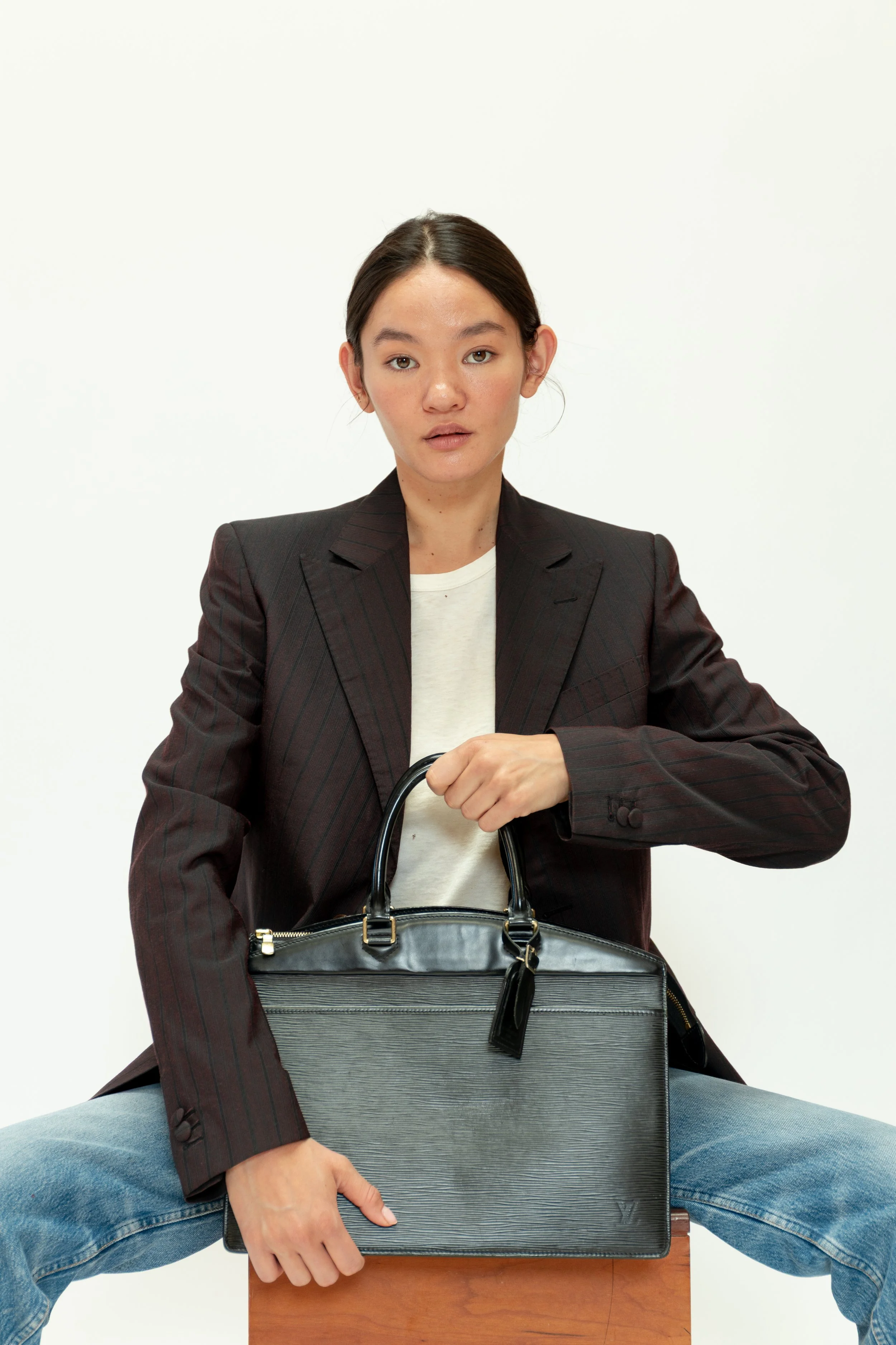 A woman sitting on a wooden box holding a black leather handbag, wearing a dark blazer and jeans, with a neutral facial expression against a plain white background.