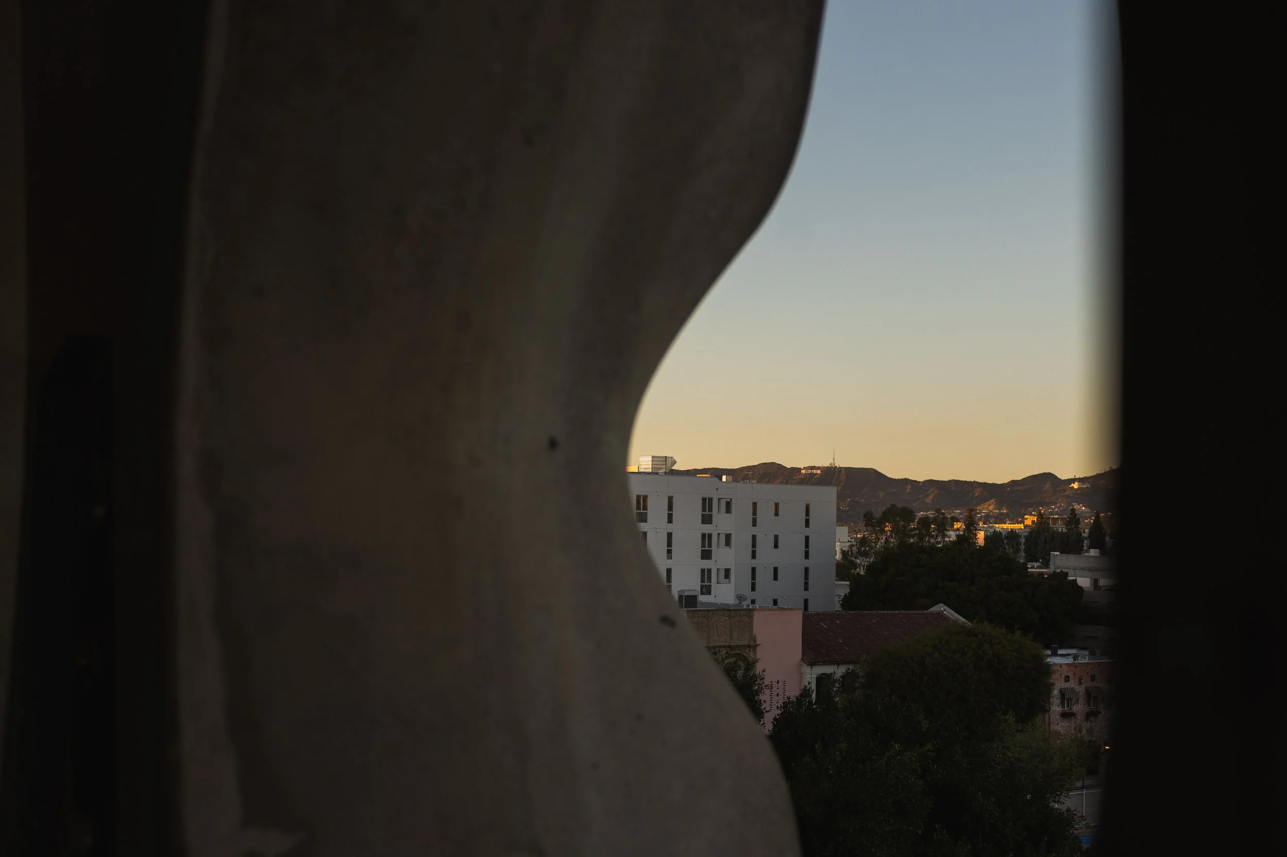 View of buildings and mountains through a rounded opening in a concrete or stone structure at sunset or sunrise.
