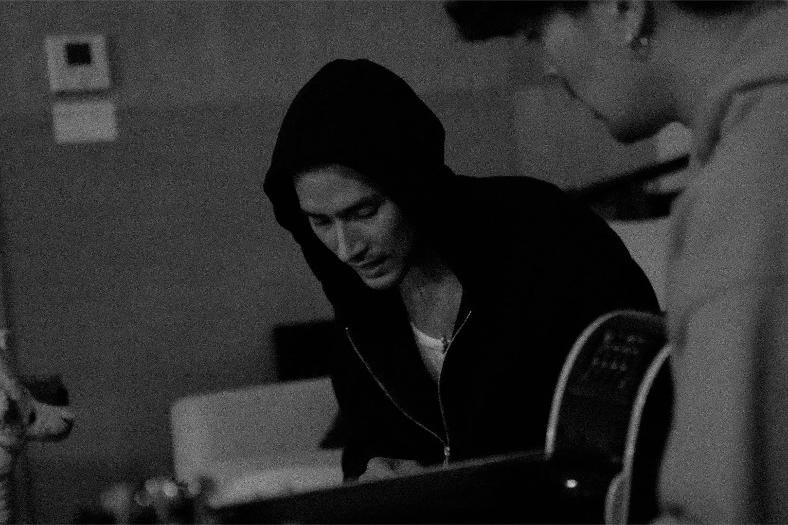 Black-and-white photo of a woman wearing a hoodie, looking down, with a man playing guitar in the foreground.