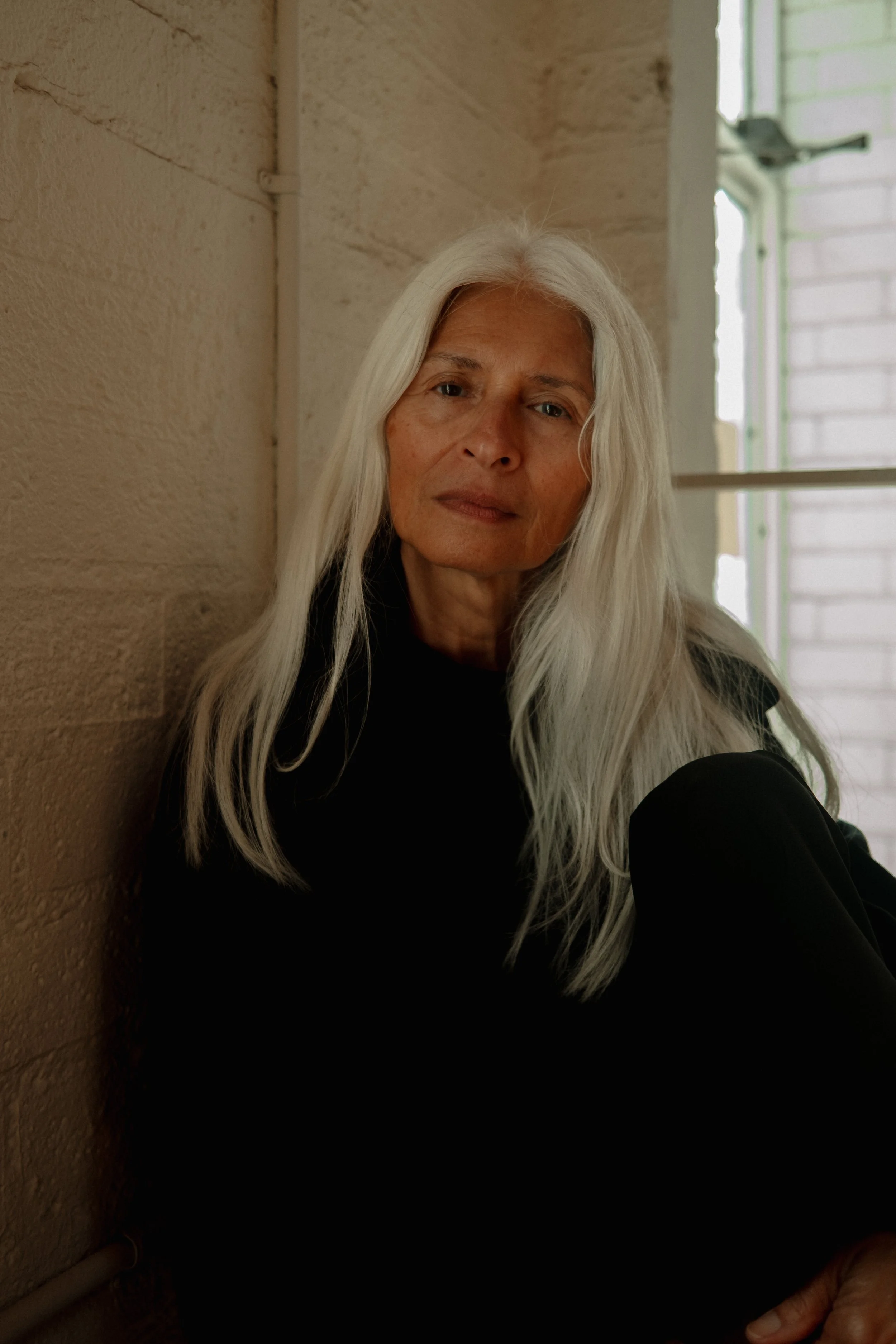 A woman with long white hair sitting against a brick wall, wearing a black top, near a window with soft natural light.