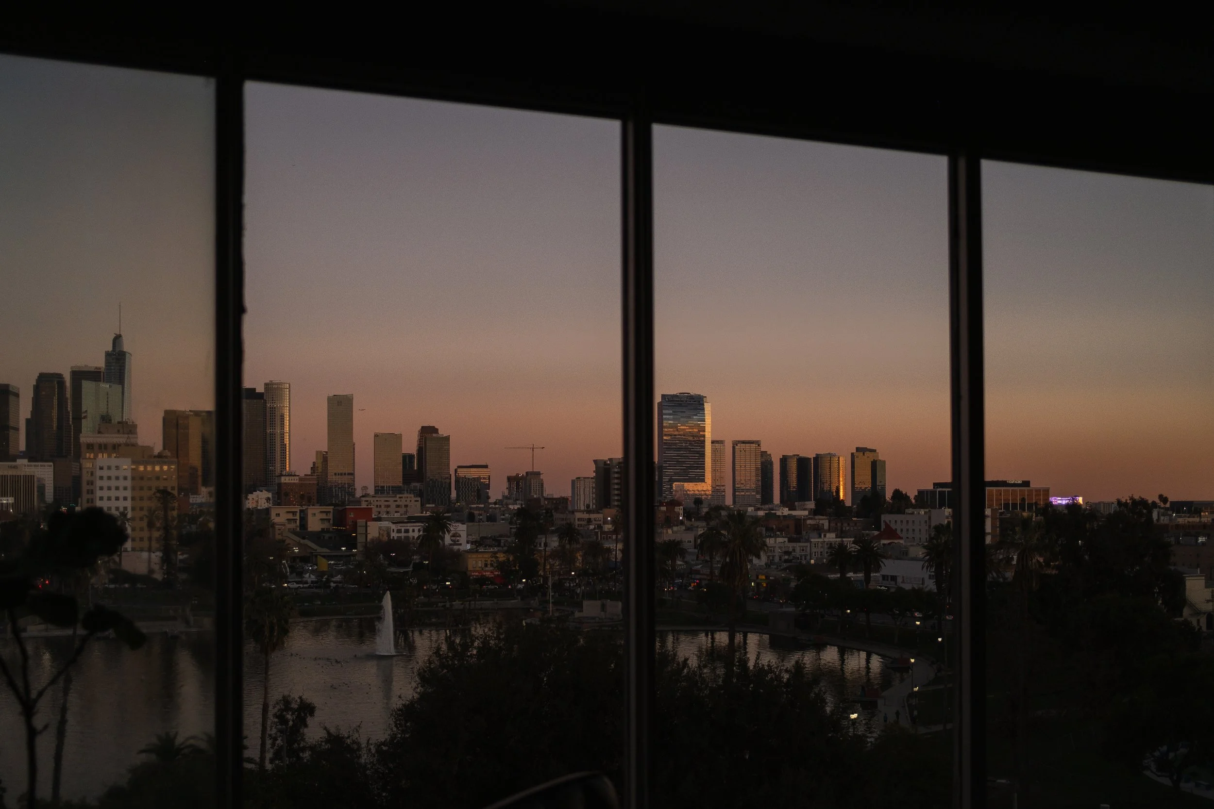 City skyline at sunset viewed through large window with framing bars, including high-rise buildings and a pond with fountain in the foreground.