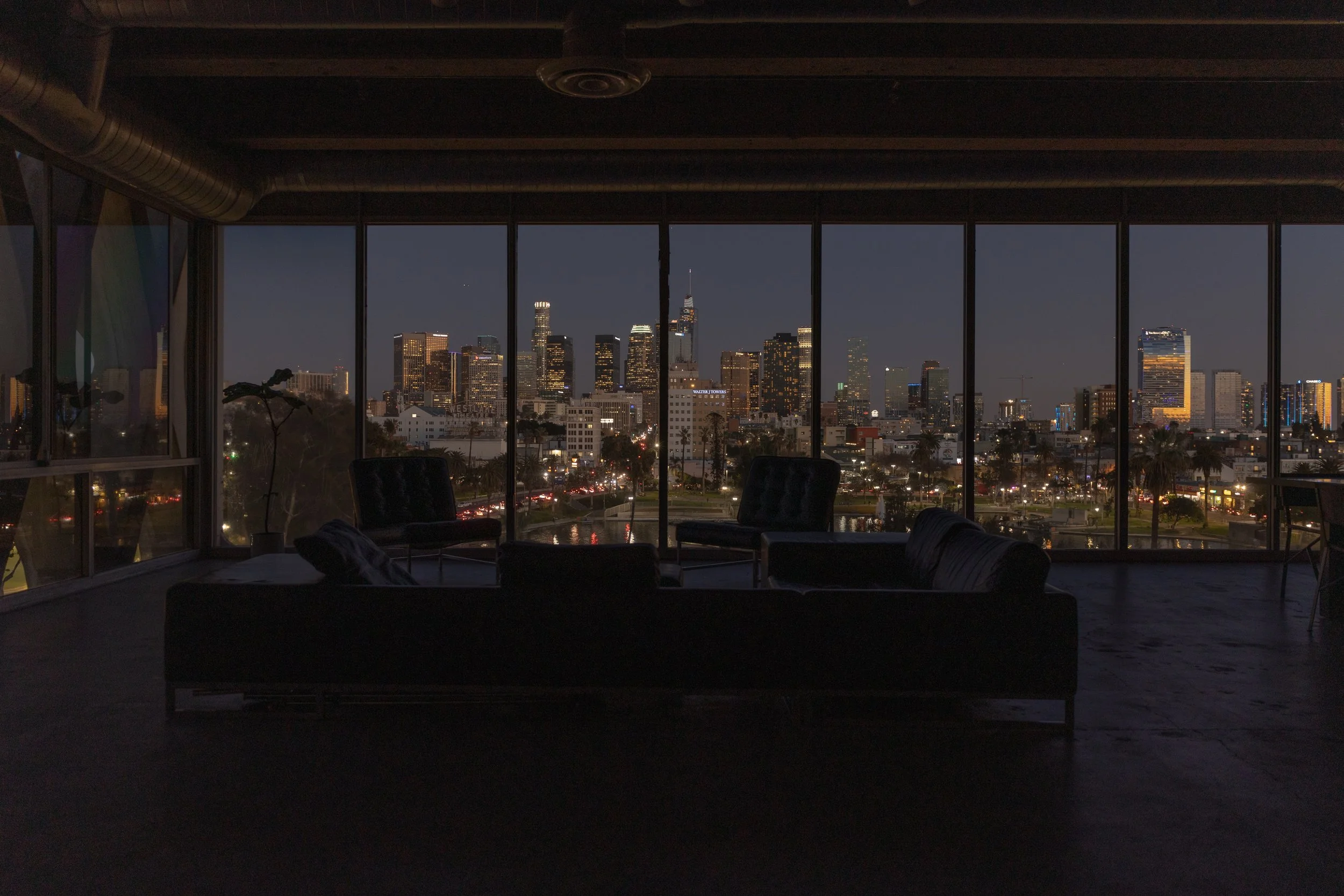 Nighttime city skyline view through large windows from a dark interior room with furniture.
