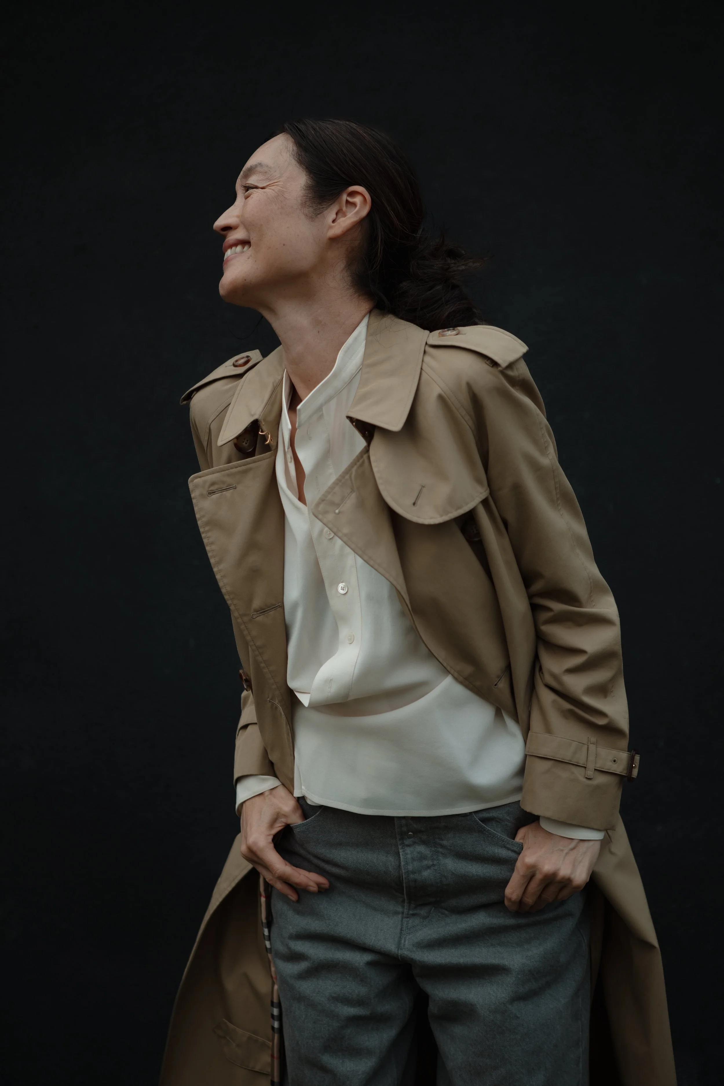 Side profile of smiling woman with dark hair wearing a beige trench coat and light-colored shirt against a dark background.