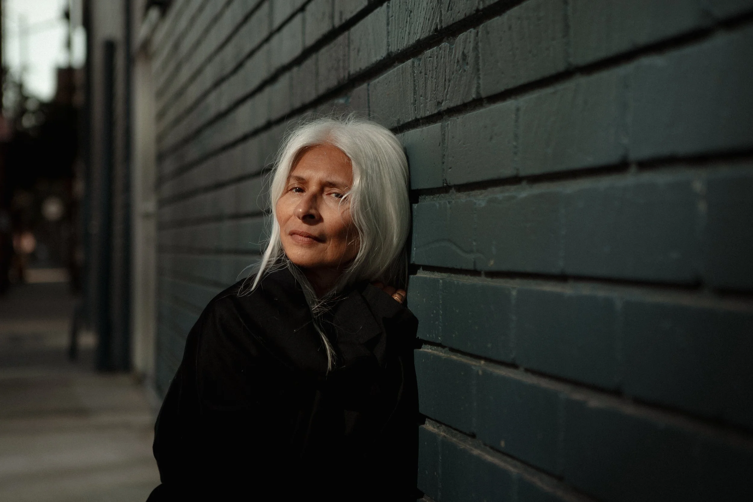 An older woman with long white hair looks at the camera while leaning against a dark green brick wall on a city street at night.