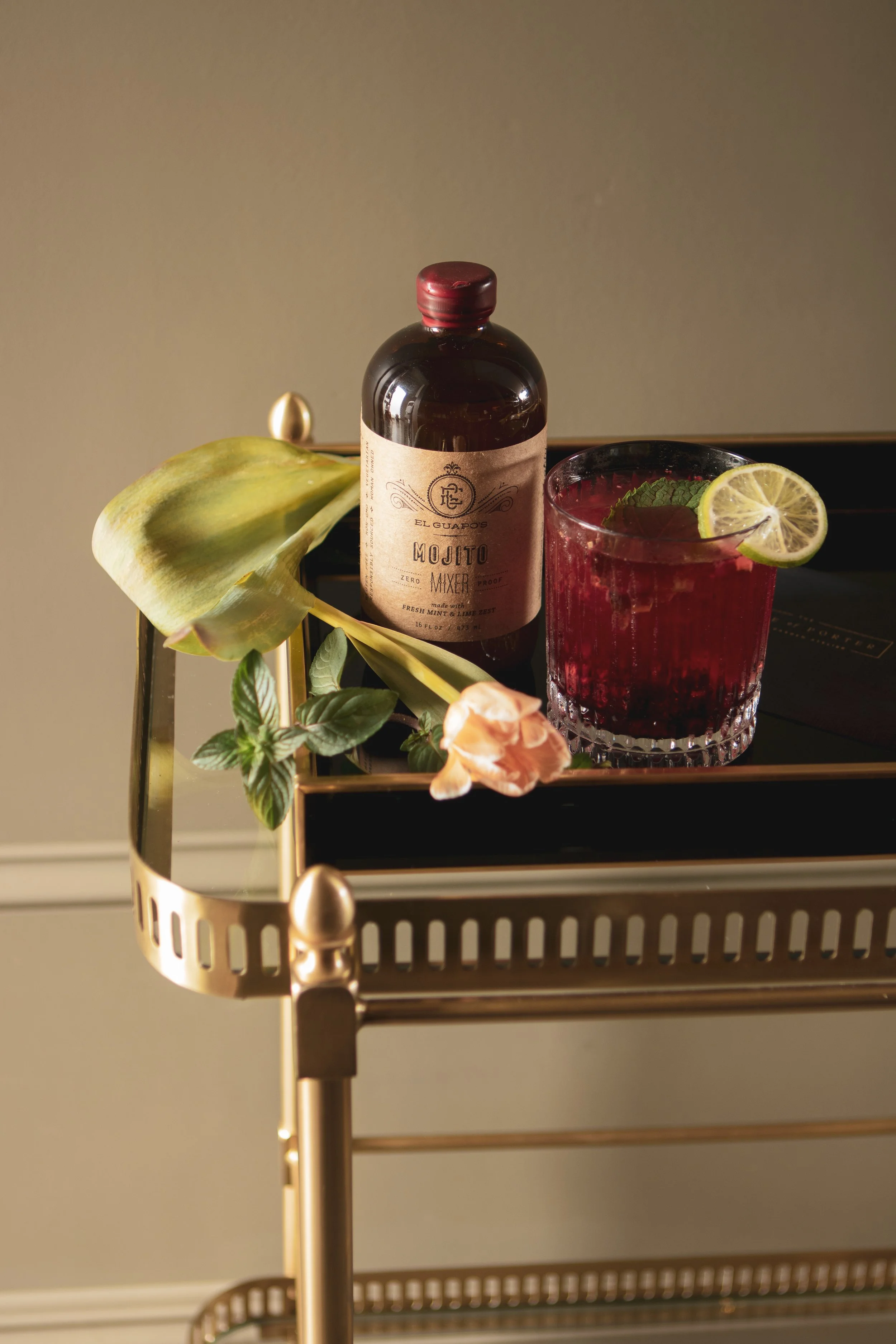 A cocktail drink garnished with a lemon slice on a glass, a bottle of El Guapo's Mojito Mixer, and decorative greenery on a gold and black serving cart.
