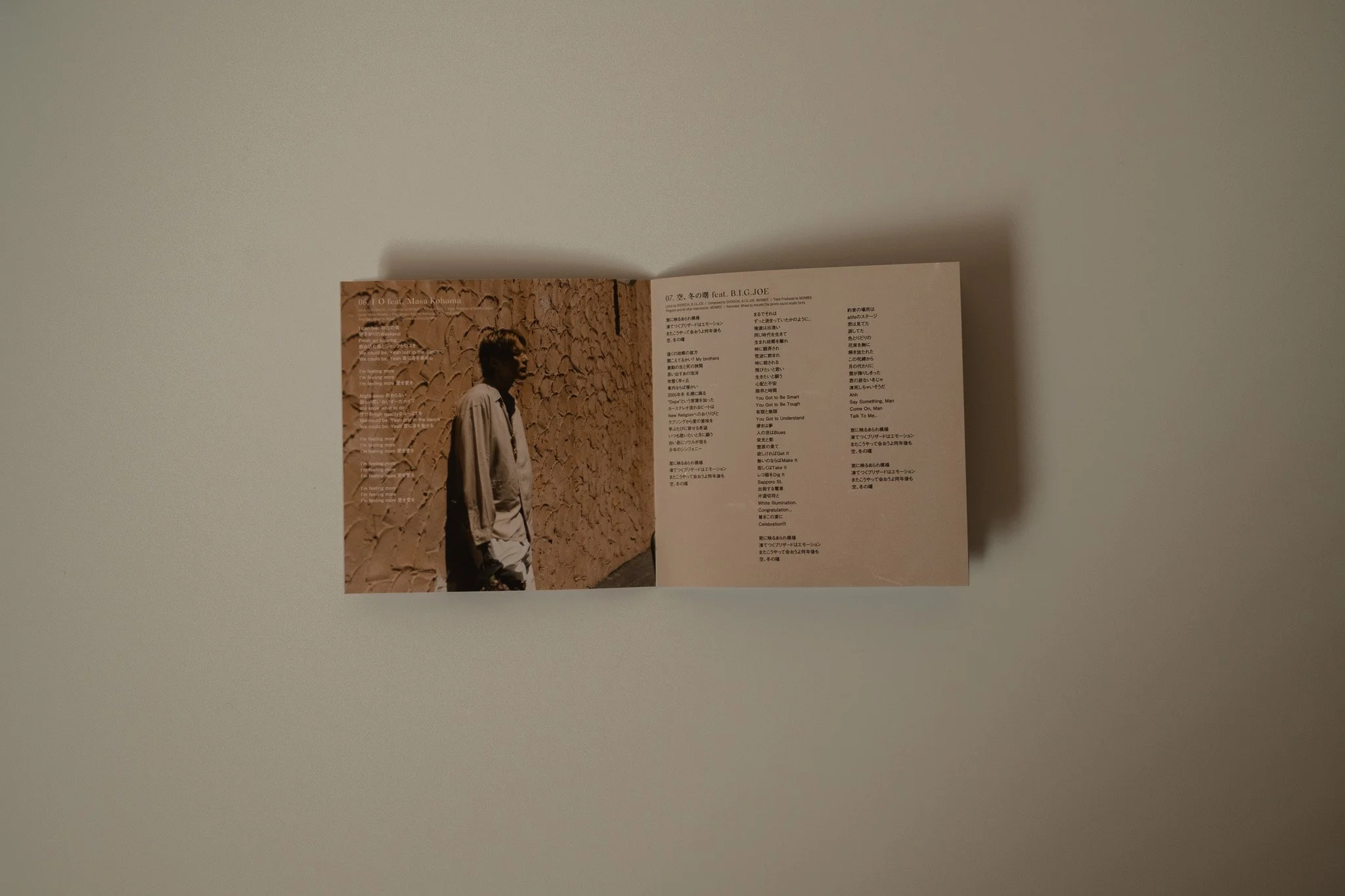 Open booklet laid flat on a white surface, featuring a photo of a man standing against a textured wall on the left page, and song lyrics in Japanese and English on the right page.