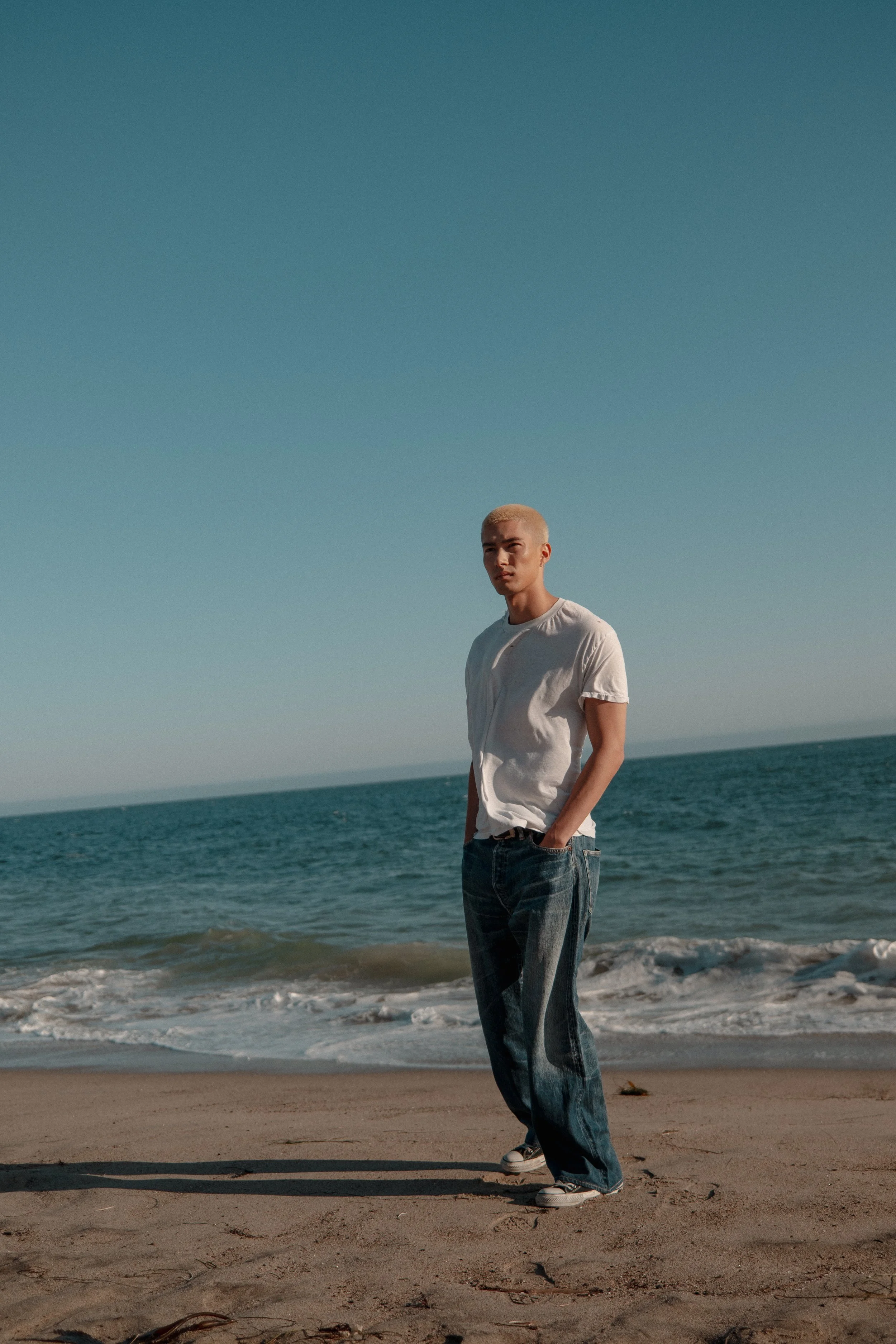 A young man with a buzz cut hairstyle wearing a white t-shirt and baggy jeans stands on the sandy beach with the ocean and clear sky in the background.
