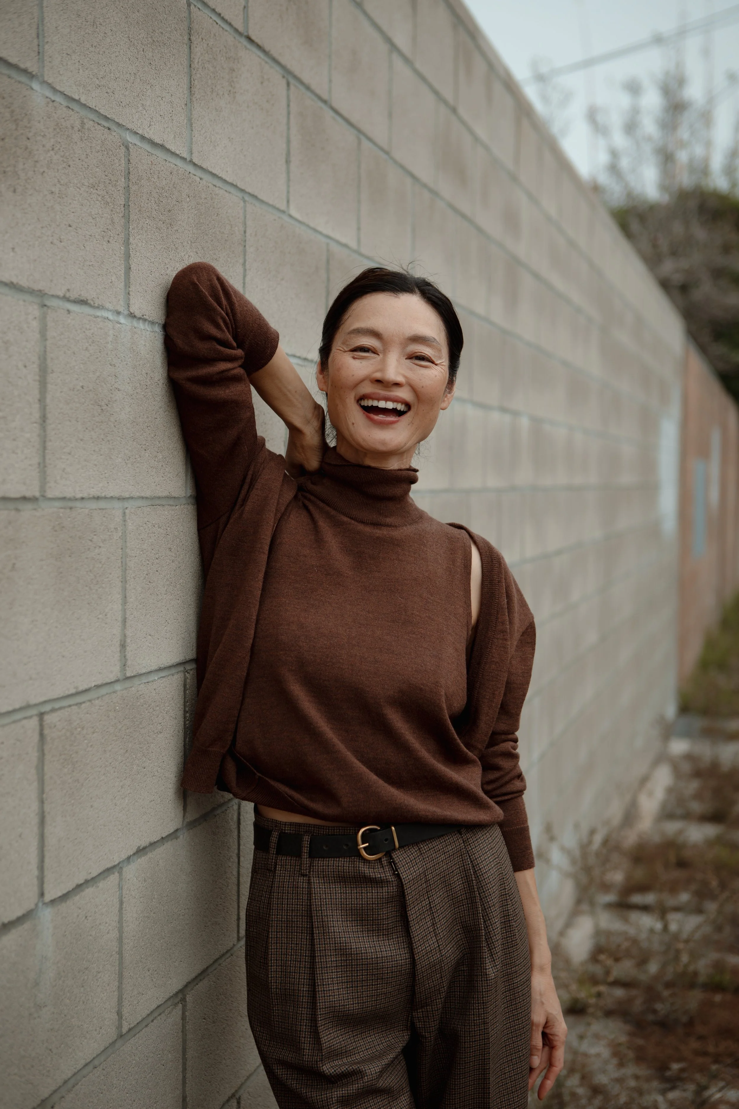 A woman with short dark hair, wearing a brown turtleneck top and plaid pants, smiling happily while leaning against a brick wall outdoors.