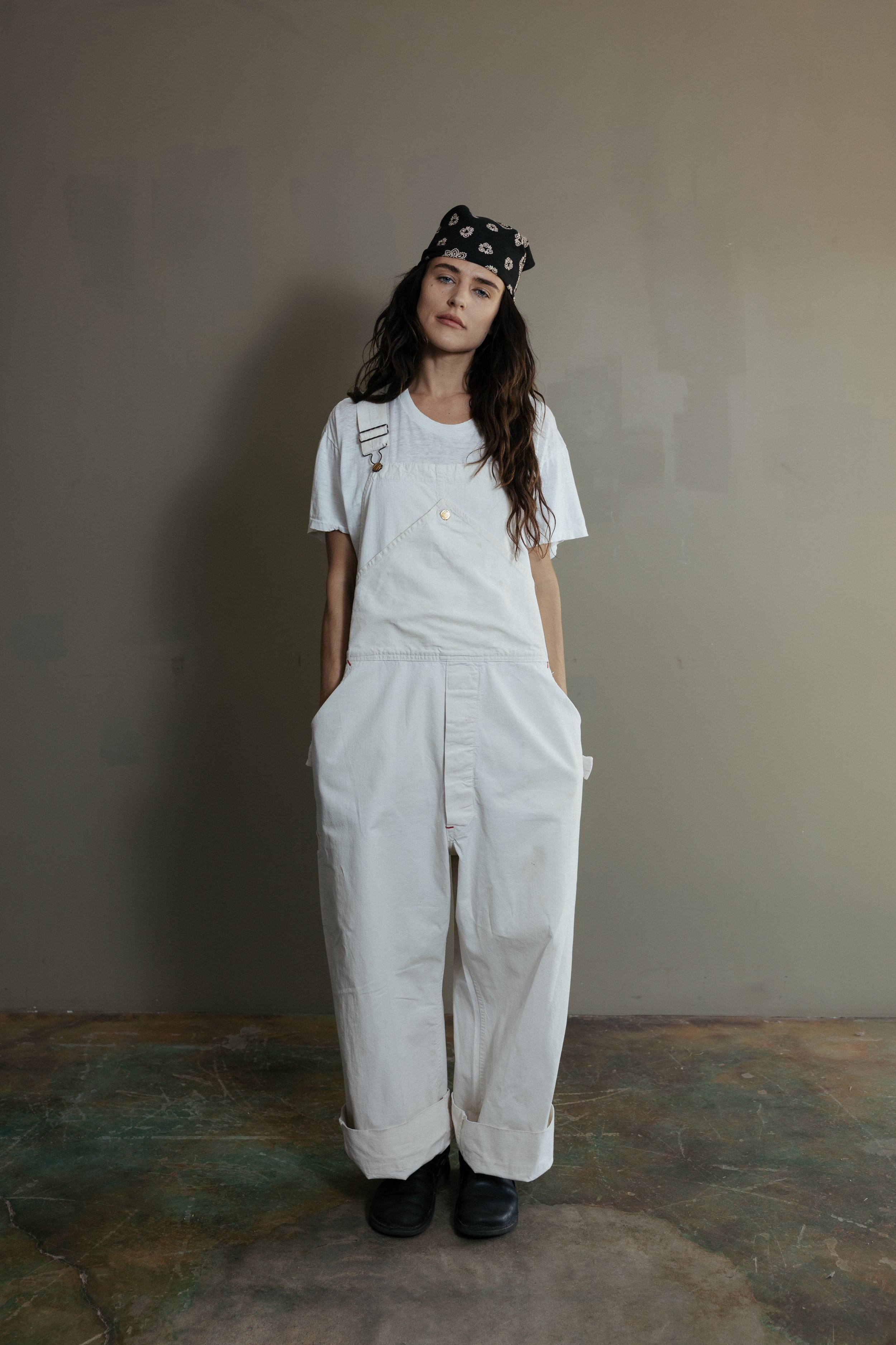 A woman standing against a plain wall, wearing a black bandana with white skull patterns, a white T-shirt, white overalls, and black boots.