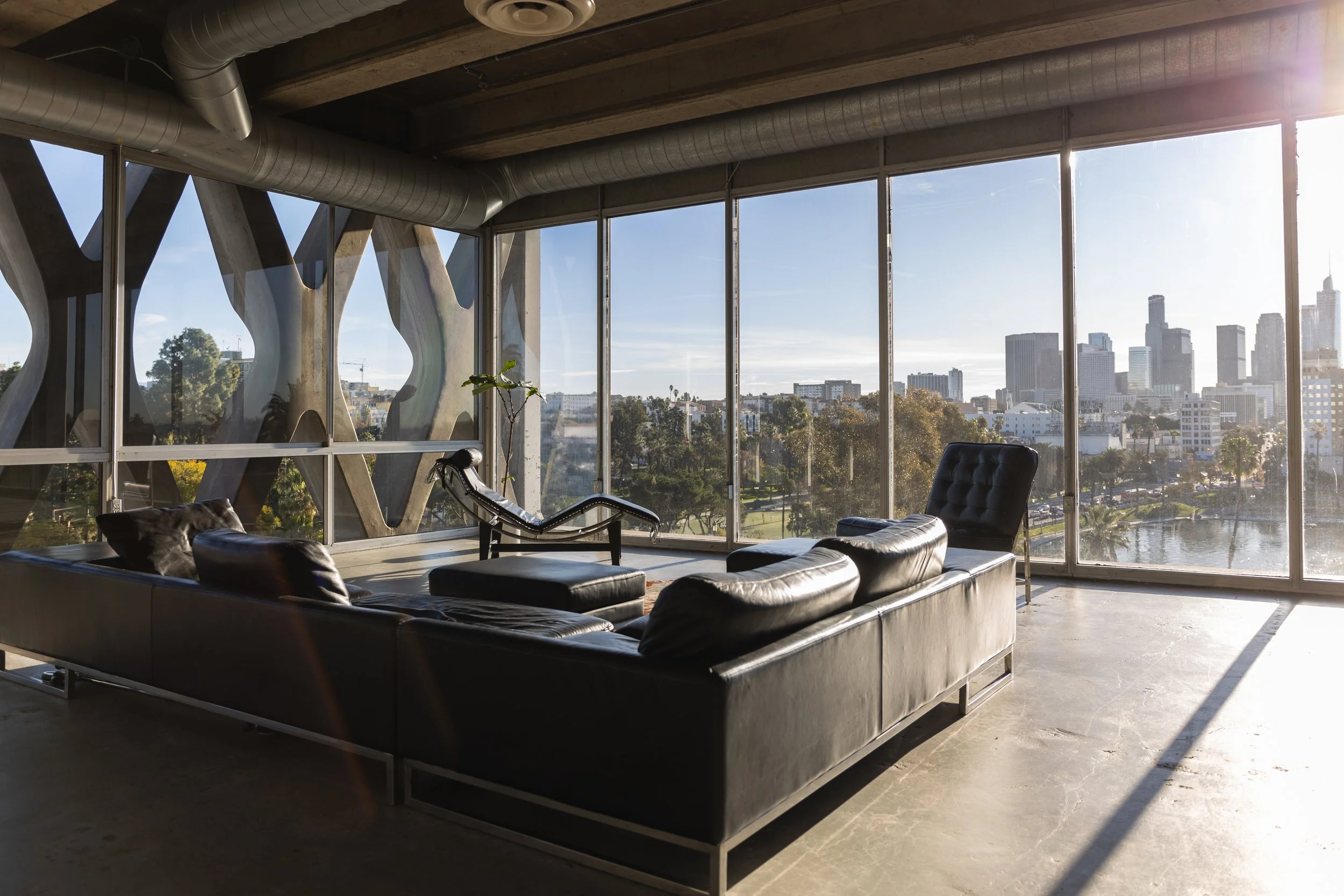Modern living room with large glass windows offering city skyline view, black leather sectional sofa, lounge chair, and a potted plant.