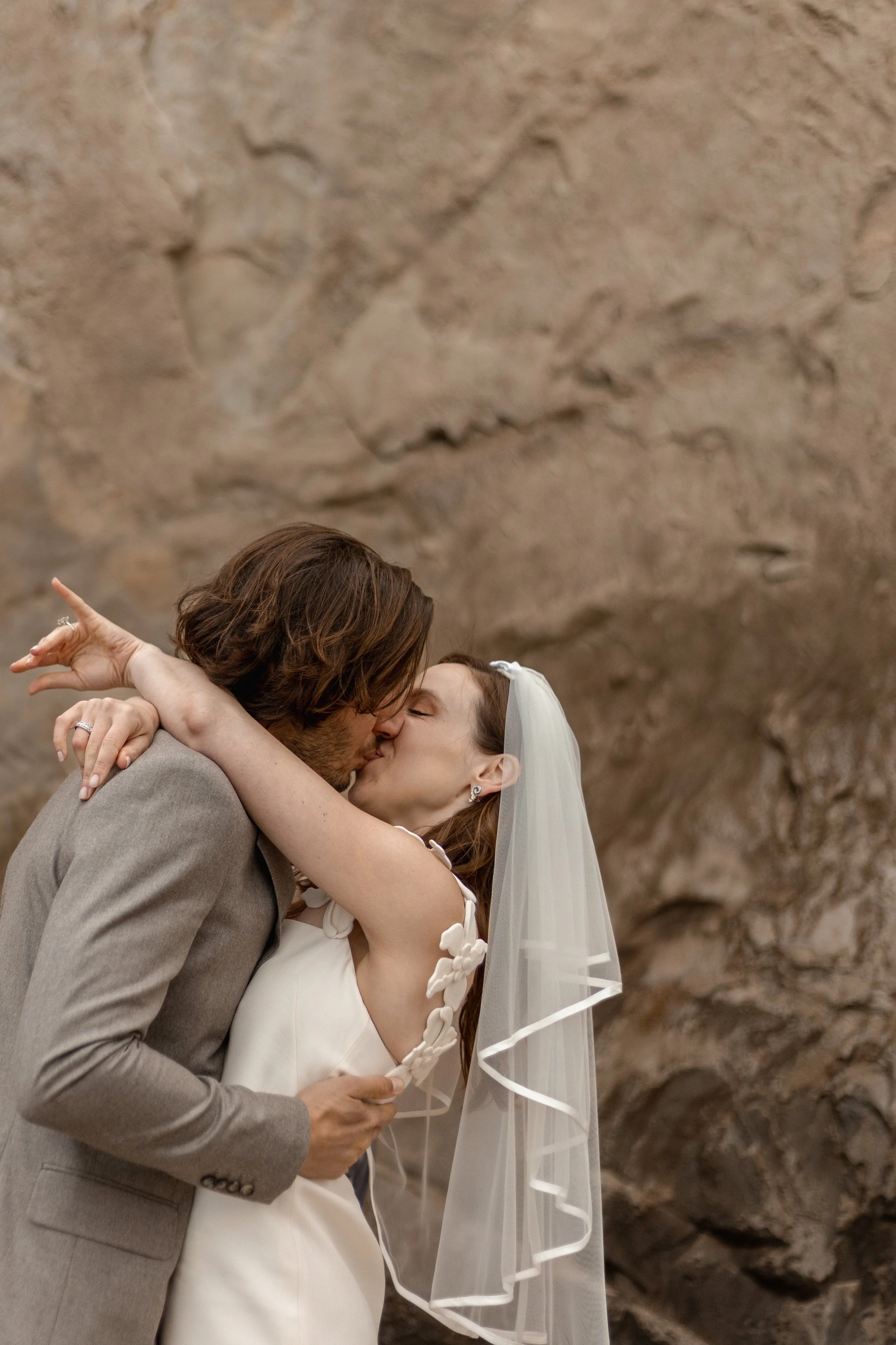 A bride and groom sharing a kiss during their wedding ceremony, with the bride wearing a veil and a white wedding dress, and the groom in a light gray suit, against a rocky background.