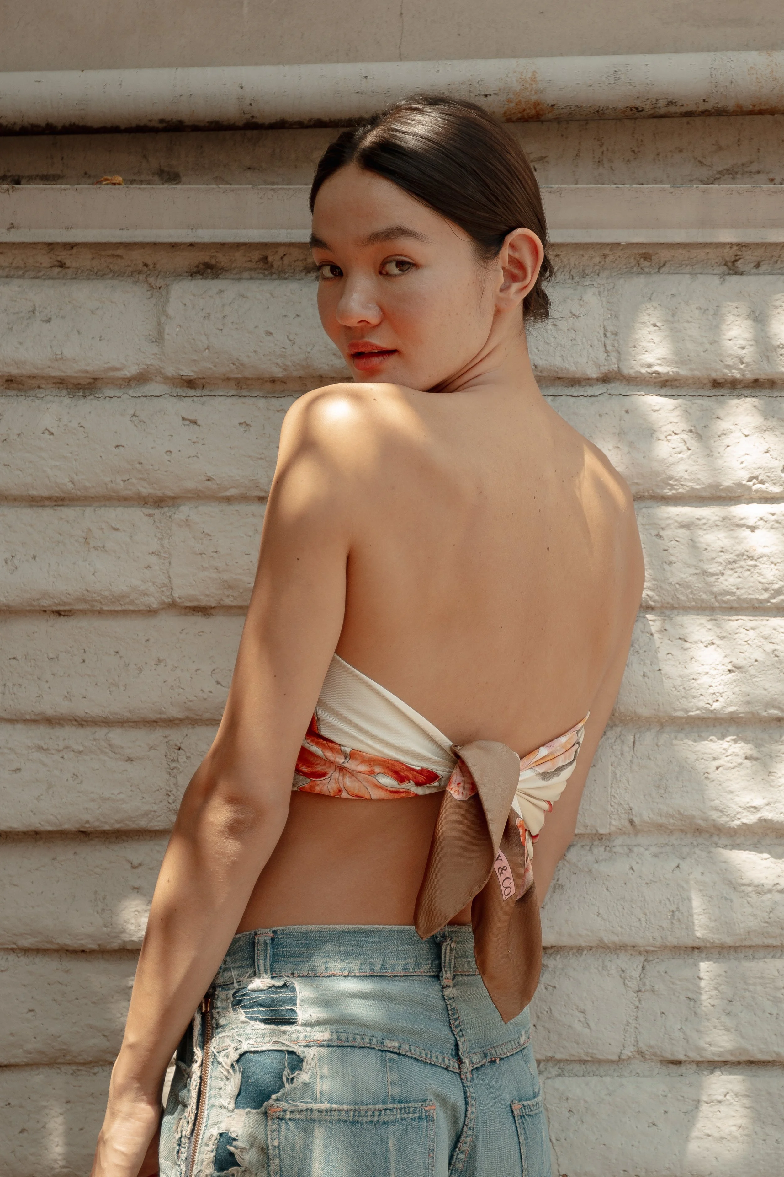 A young woman with dark hair, facing away but looking over her shoulder, standing against a textured white brick wall with shadows.