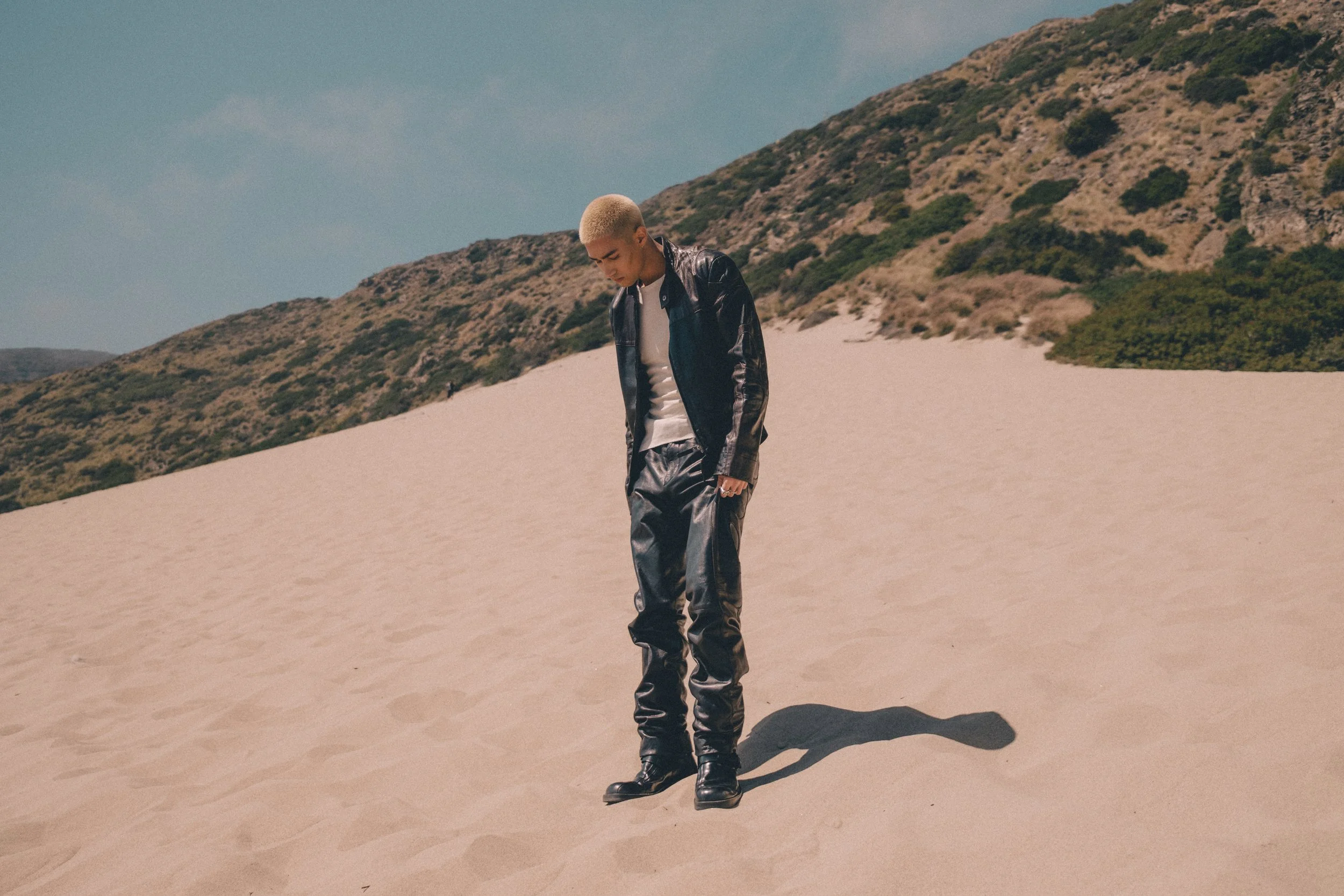 Person with blonde hair wearing a black leather jacket and black leather pants walking on sand dunes in a desert landscape.