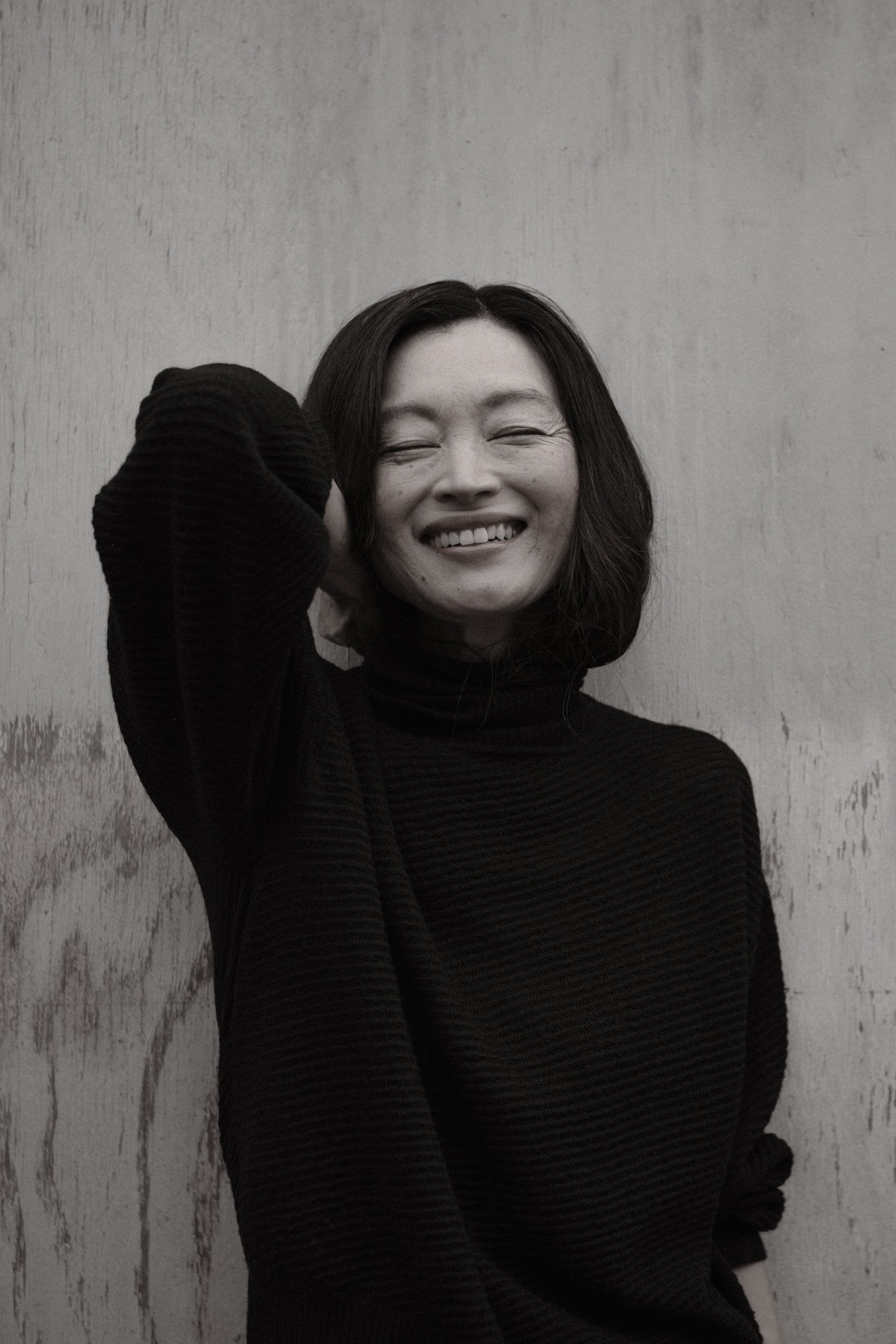 A woman with shoulder-length dark hair smiling and touching her head, wearing a black long-sleeve top, standing against a textured wall.