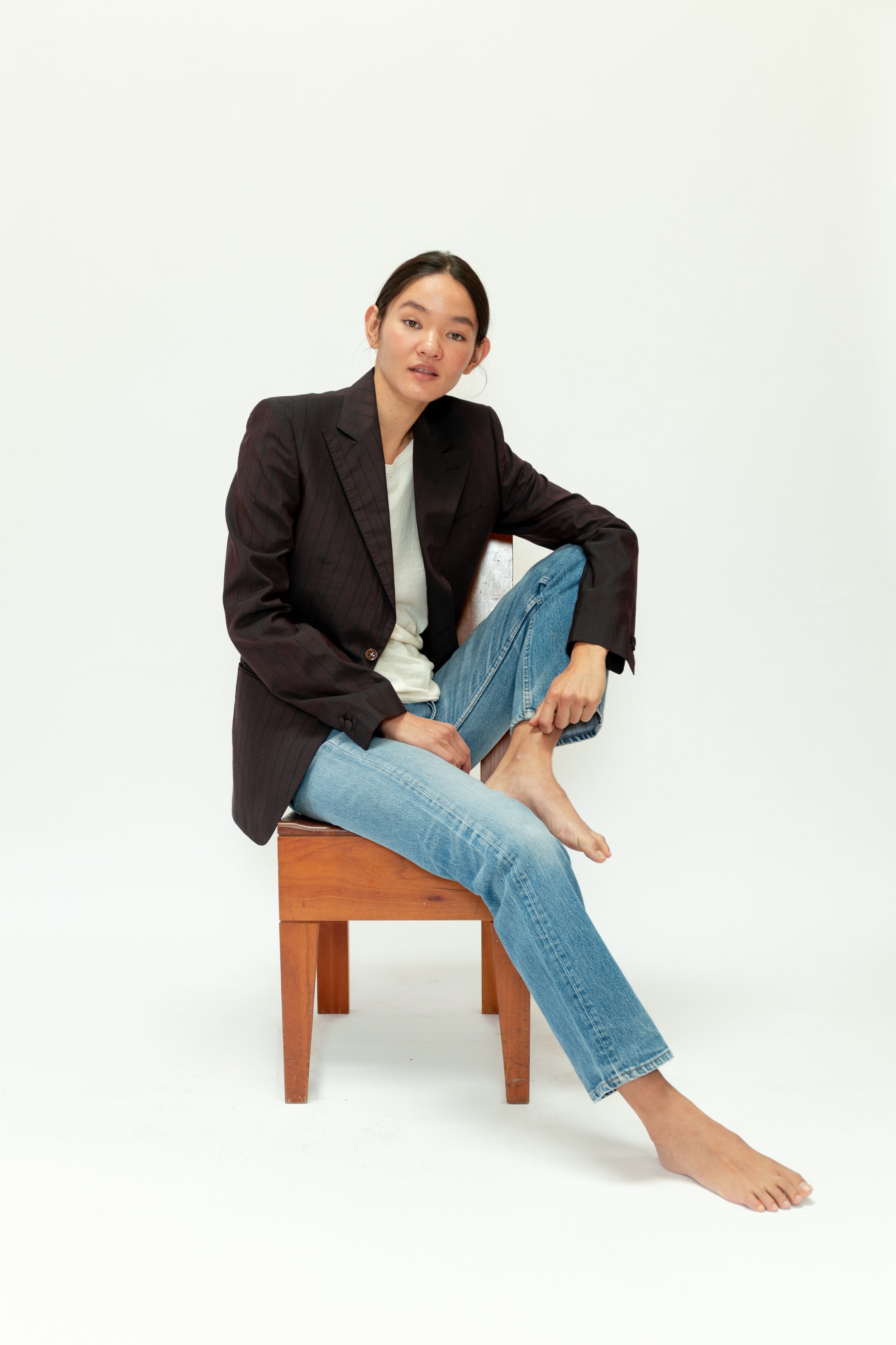 A woman sitting on a wooden stool, wearing a black blazer, a white shirt, and light blue jeans. She is barefoot and posing against a plain white background.