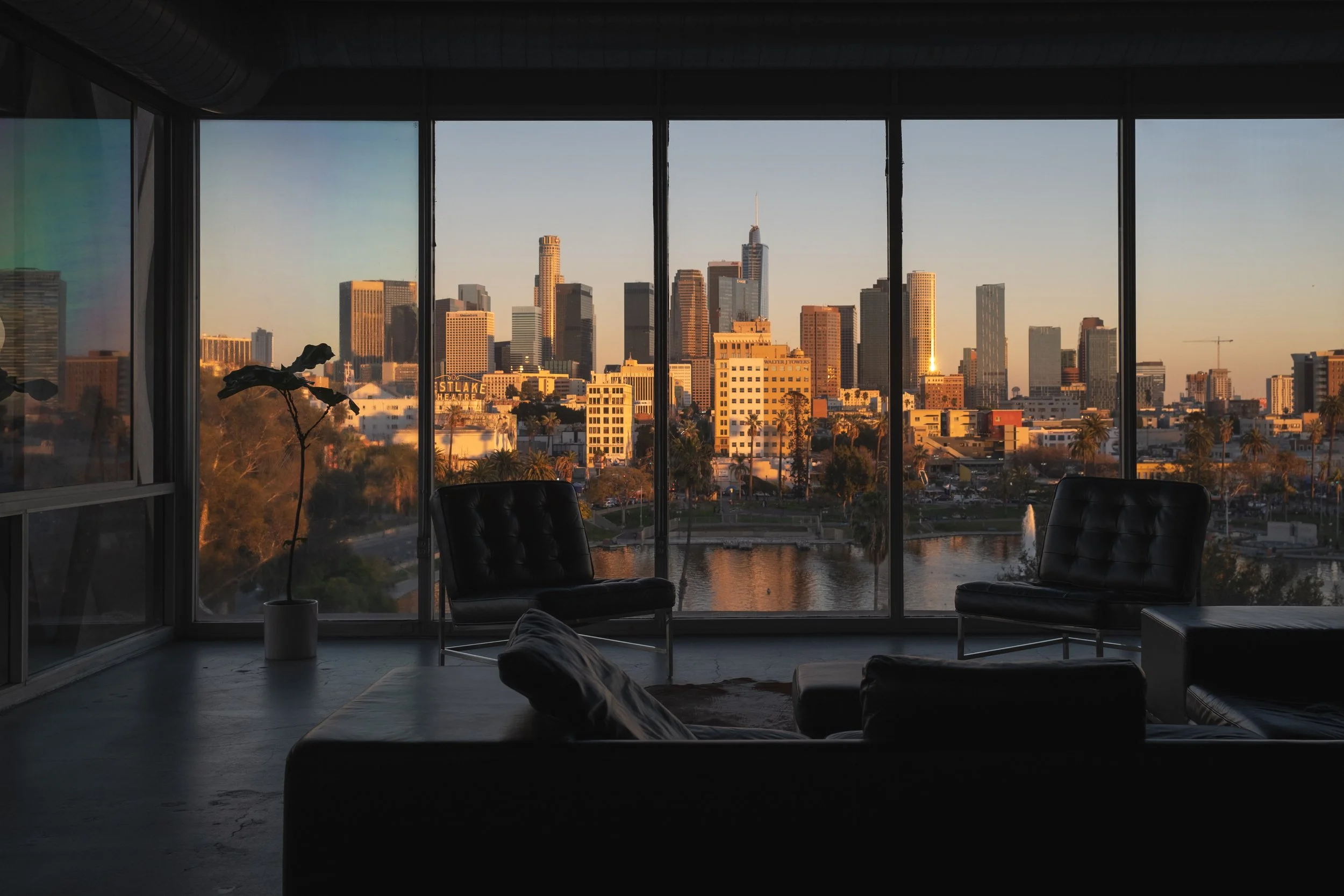 Interior of a modern apartment with large floor-to-ceiling windows showing a city skyline at sunset, with chairs and a sofa inside.