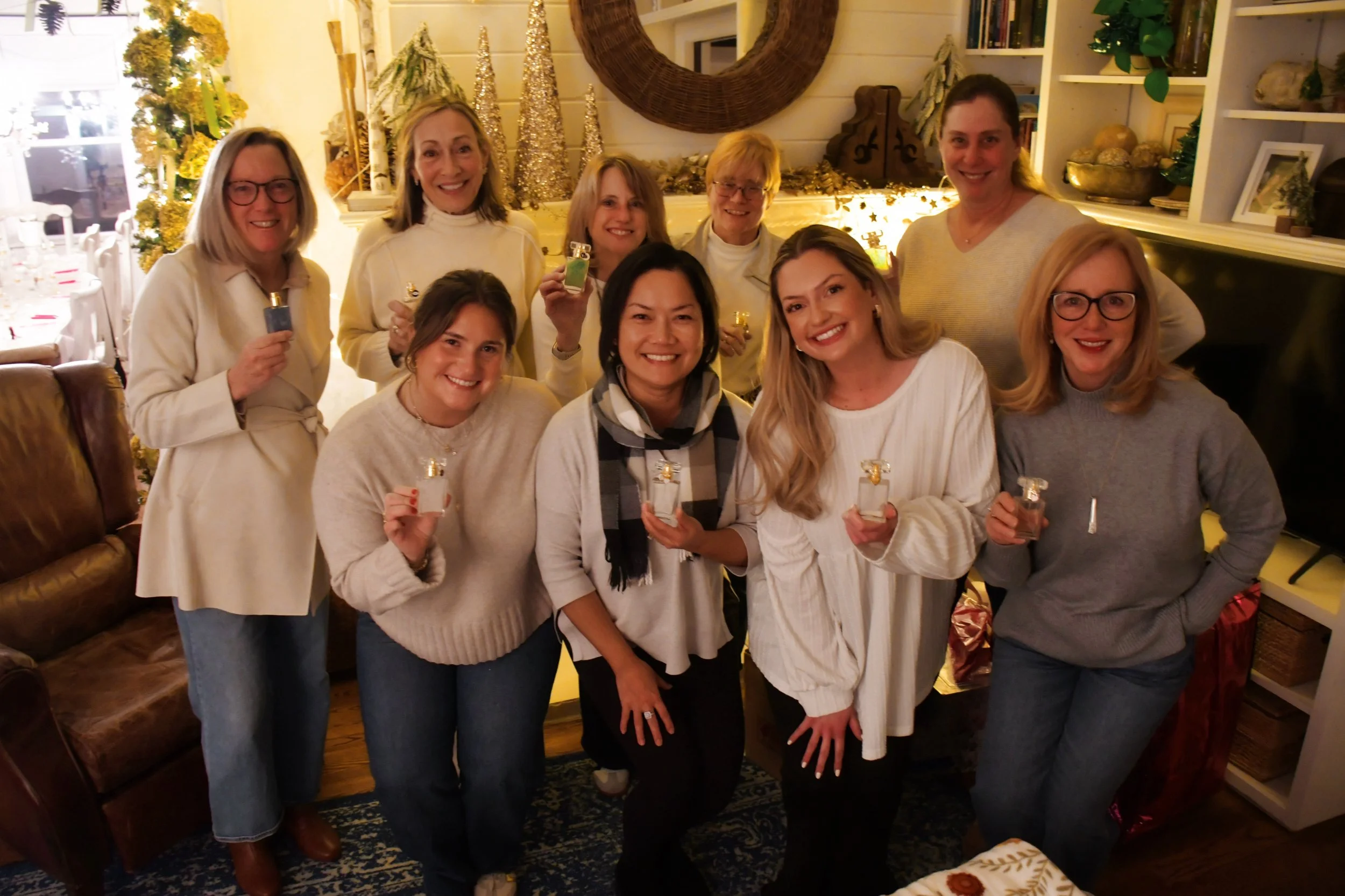 A group of ten women gathered in a cozy, festively decorated living room, smiling at the camera while holding small bottles of perfume.