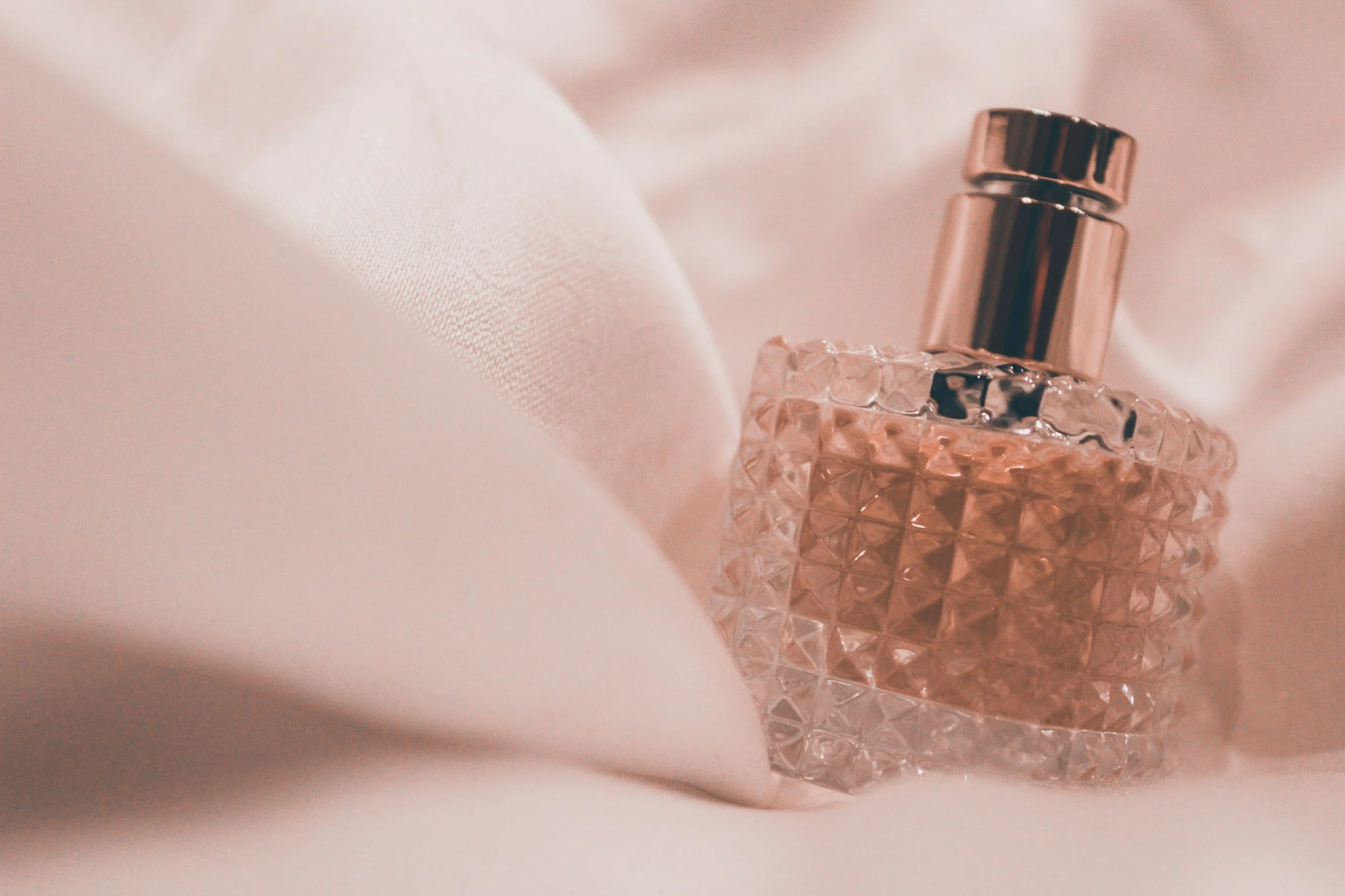 A glass perfume bottle with a textured, diamond pattern surface and a metallic cap, sitting on soft, light-colored fabric.
