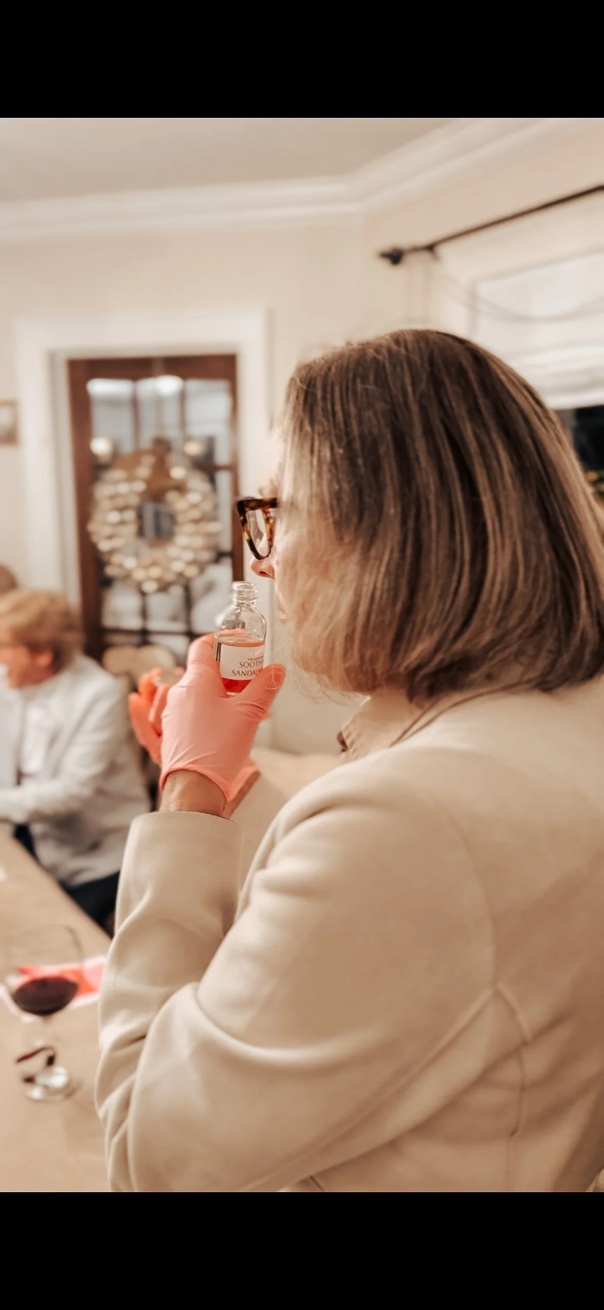 A woman with brown hair, glasses, and beige blazer is holding a small bottle near her nose in a warmly lit room, during a custom perfume making workshop