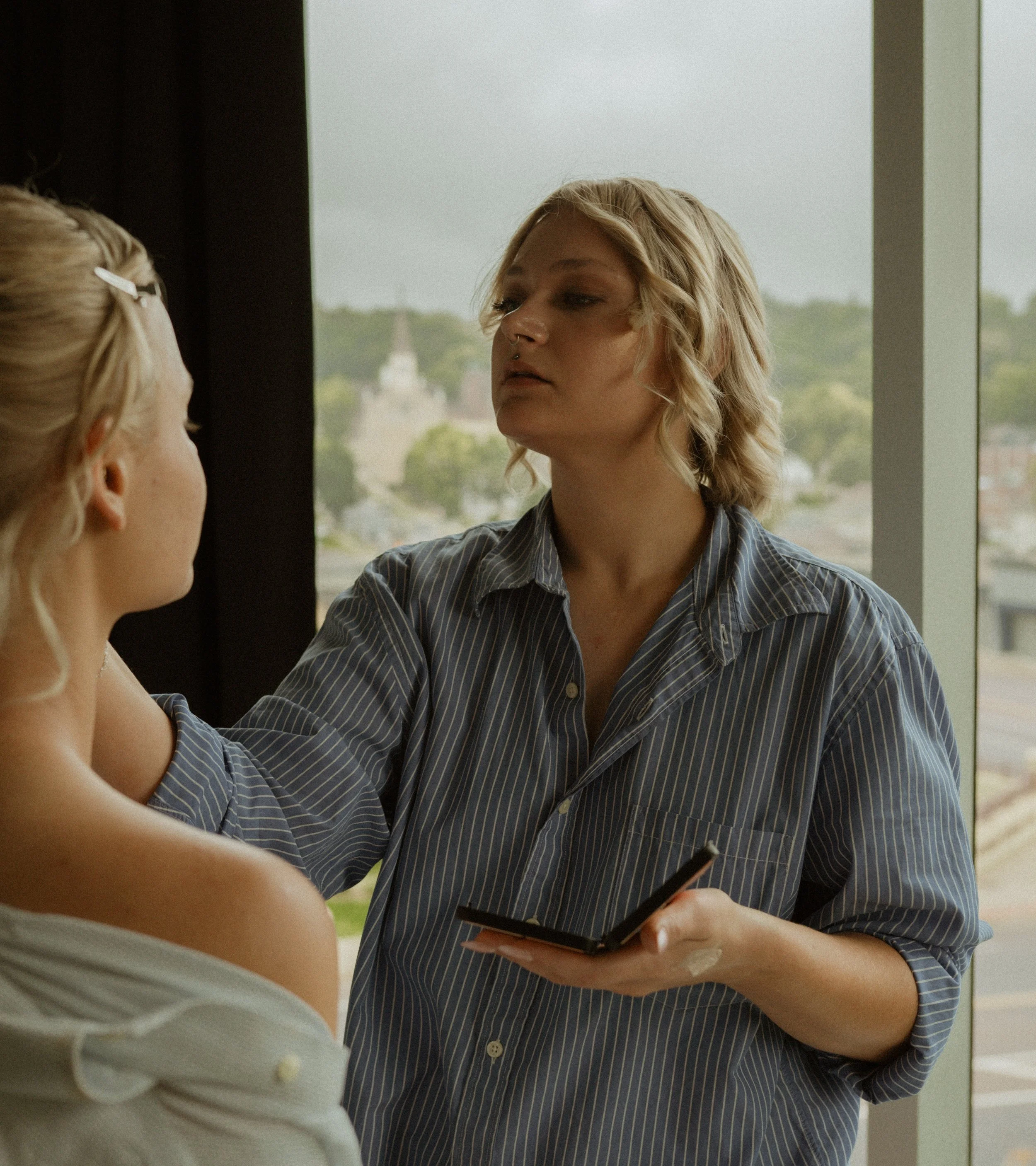 A woman with blonde hair and a nose ring holds a makeup brush while applying makeup to another woman with blonde hair tied back wearing an off-the-shoulder white top, near a large window showing a cityscape with trees and buildings in the background.