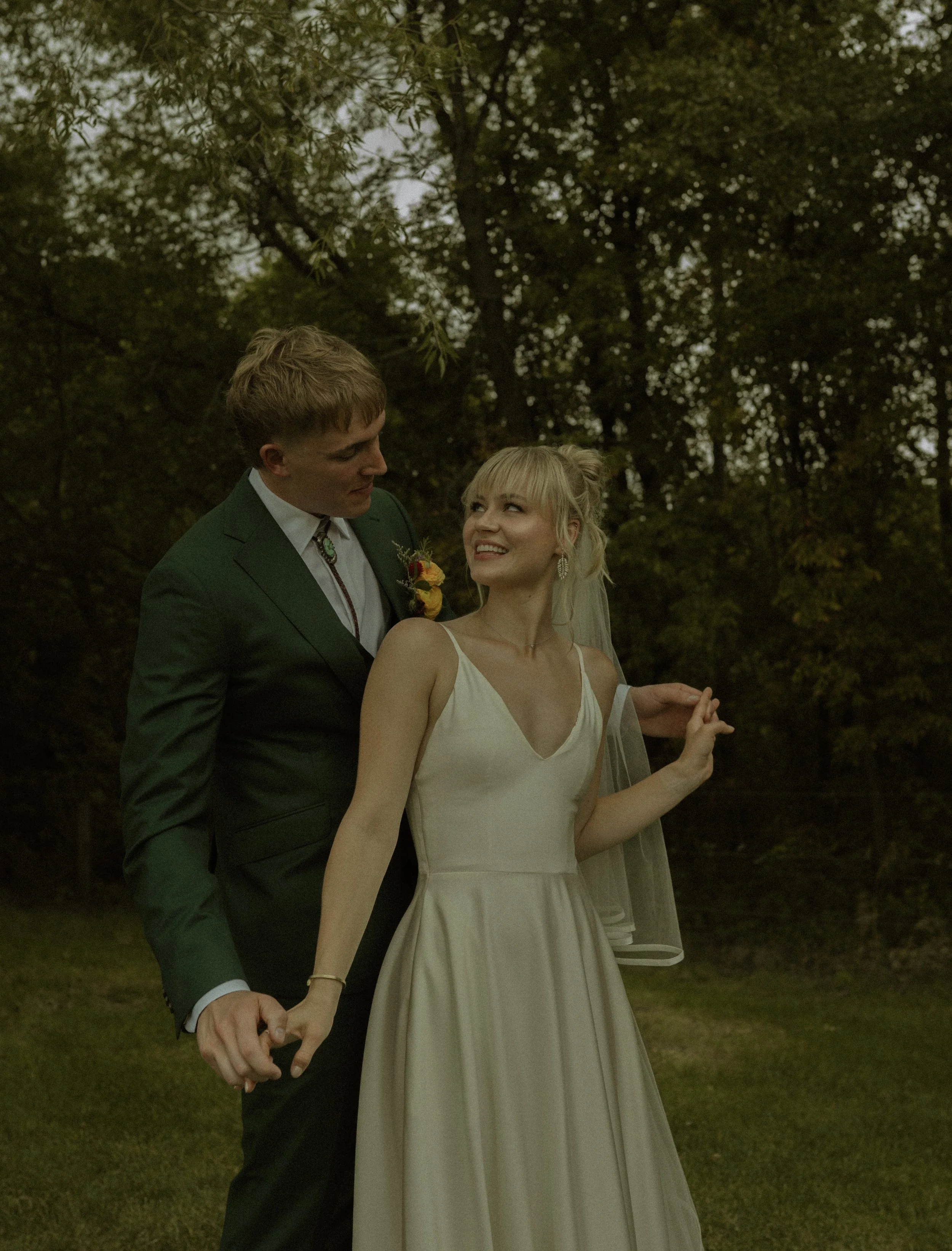 A smiling bride and groom holding hands outdoors with trees in the background, celebrating their wedding.
