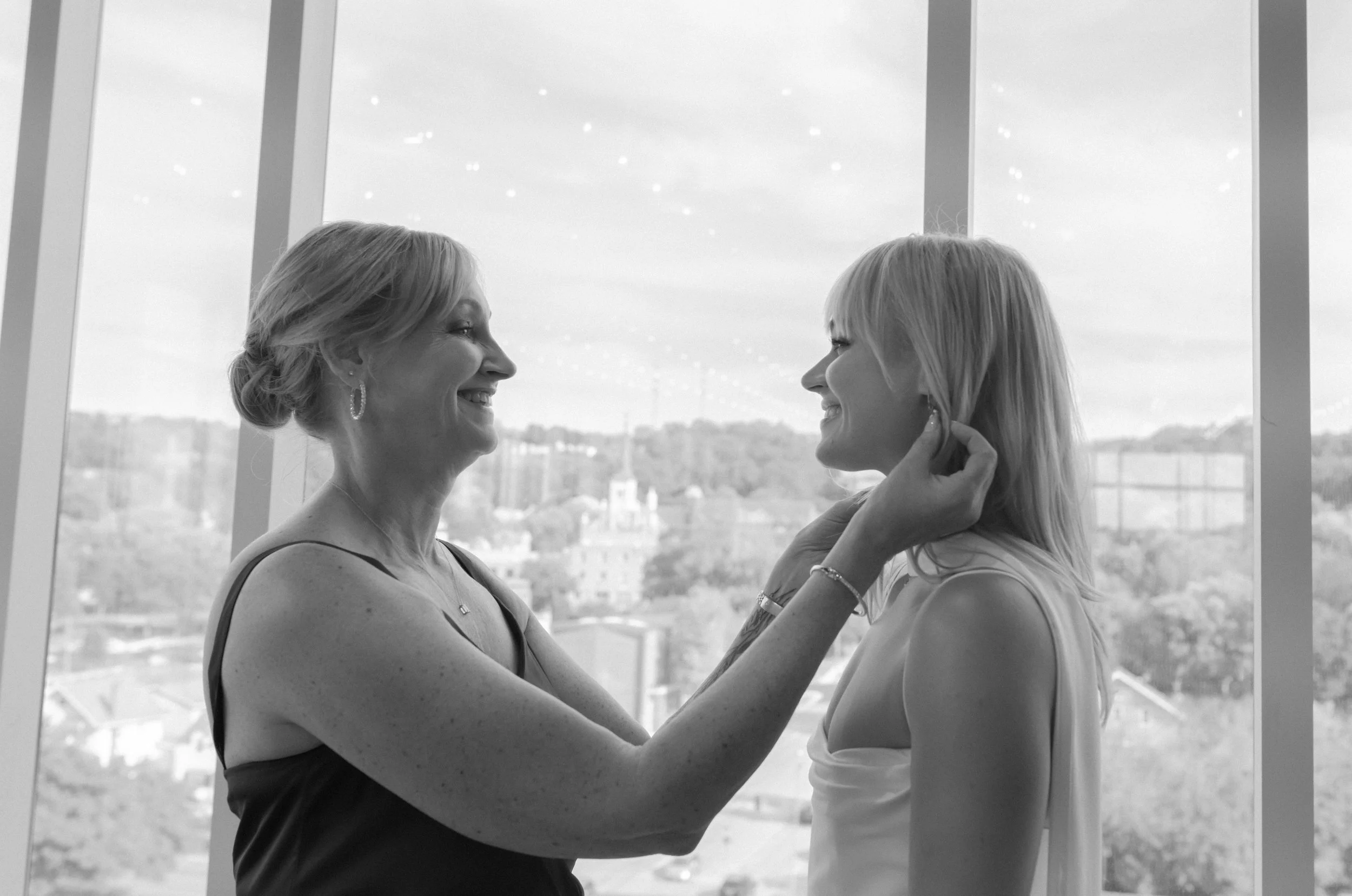 Two women smiling at each other, one helping the other with earrings, standing in front of large window overlooking a cityscape.