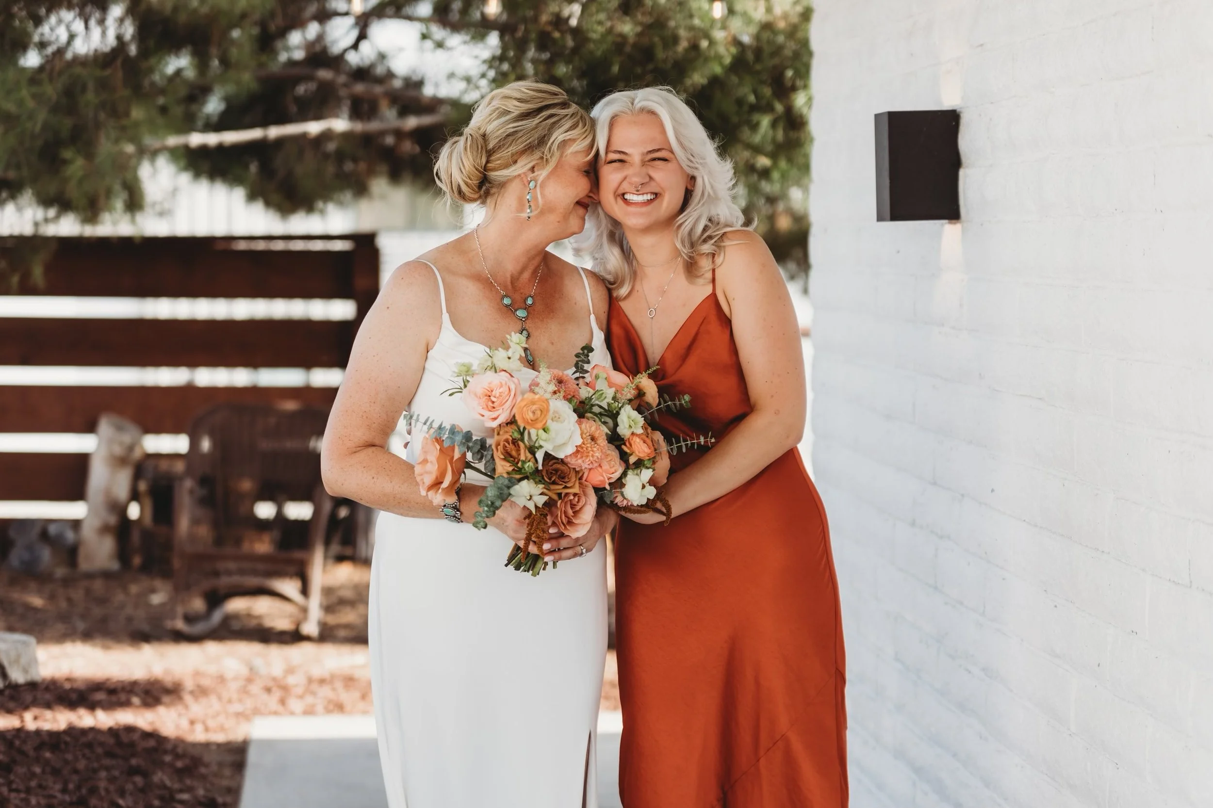 A joyful woman with gray hair in a rust-colored dress holds a bouquet of pink, white, and peach flowers, standing next to an older woman with blonde hair in a white dress with thin straps, sharing a moment of affection outdoors near a white brick wall and trees.