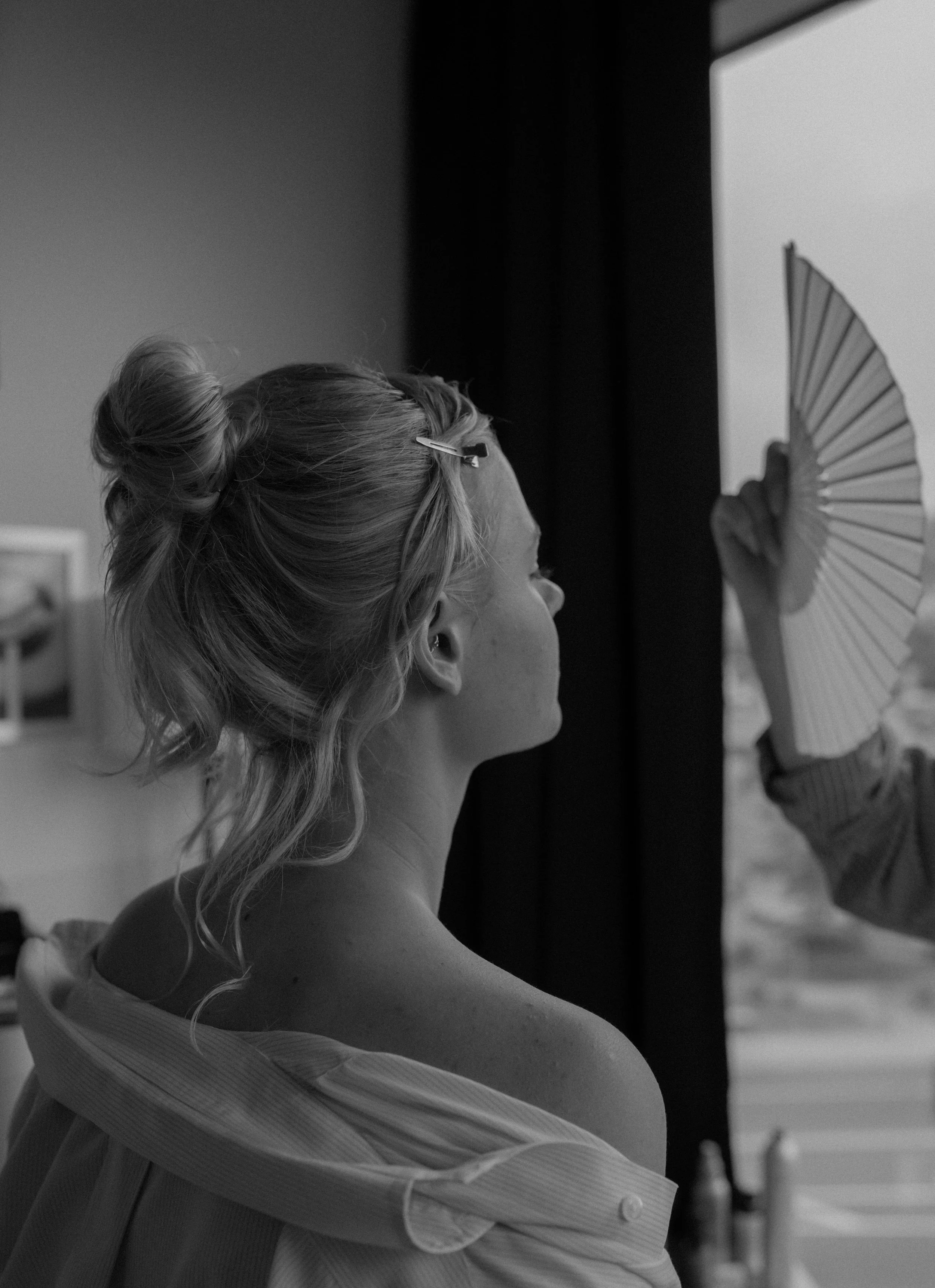 A woman with blonde hair styled in a loose bun with a hair clip, sitting with her shoulders exposed, as a person beside her holds a folding fan near her face during a makeup or hair styling session.