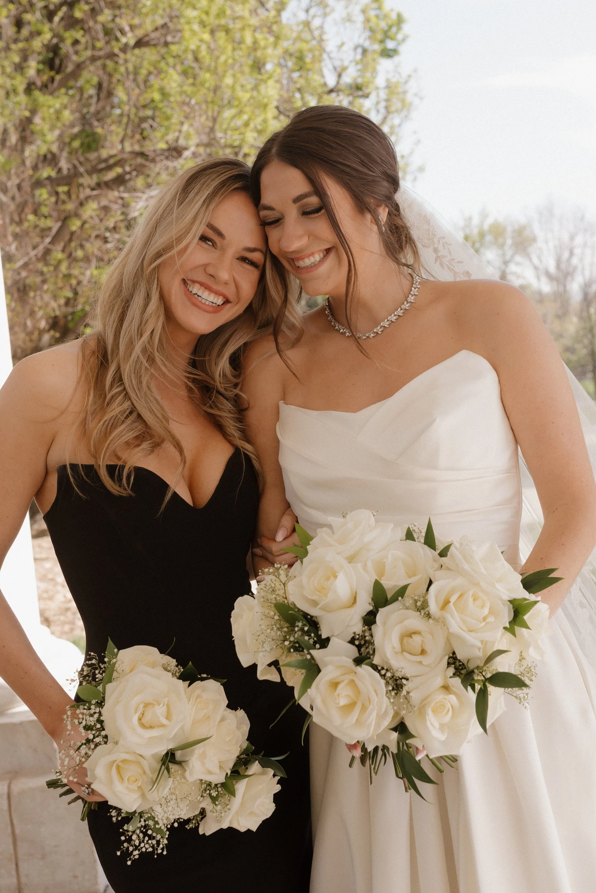 Two women smiling and leaning close together, one in a white wedding dress holding a bouquet of white roses, and the other in a black dress holding a smaller bouquet, at a wedding celebration outdoors with trees in the background.